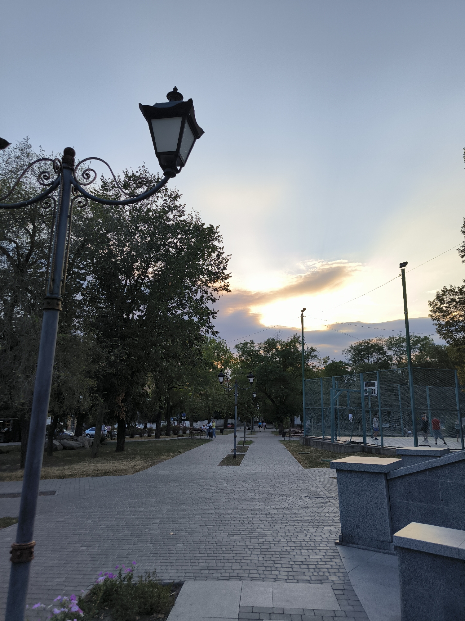 This image depicts a park path in Mykolaiv, Ukraine, with a classic streetlamp in the foreground. The path is paved with grey interlocking bricks and curves to the right as it recedes into the distance, lined by trees. On the right side of the path, a fenced basketball court is visible with several individuals playing. In the background, more trees and a soft sunset sky with clouds add to the serene atmosphere. The streetlamp in the foreground has an ornate black metal design with a white glass lantern.