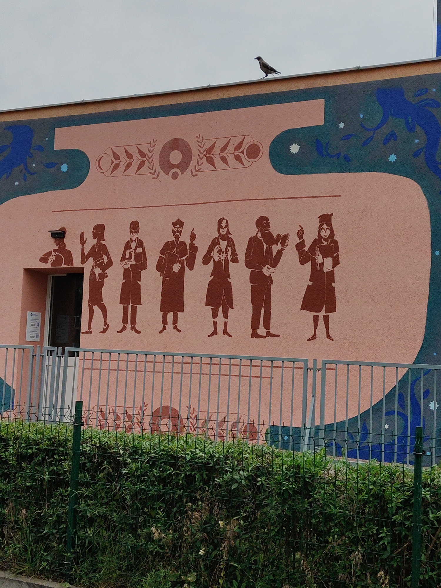 The image shows a mural on the side of a building, depicting a row of silhouetted figures in brown. The figures appear to be dressed in historical or academic attire, with some holding books or instruments. The mural has a pinkish-orange background, and to the left of the figures, there is a dark doorway with a white door and a sign. Above the mural, there is a dark bird standing on the edge of the roof. The building is partially obscured by a green metal fence and lush green bushes in the foreground. The sky is overcast and gray.