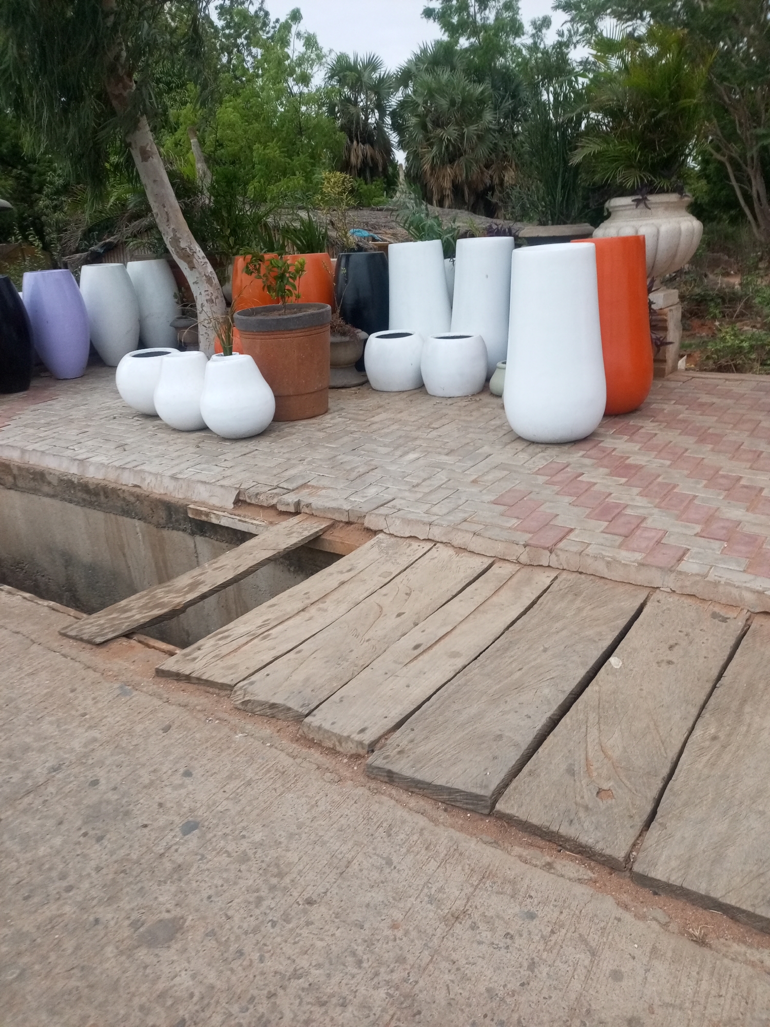 The image depicts an outdoor display of various planters, situated beside a shallow trench covered by wooden planks. The planters are a mix of shapes, sizes, and colors including black, lavender, white, and orange. They are arranged on a tiled surface that transitions into a concrete area where the trench and wooden planks are located. In the background, greenery suggests a garden or natural area. The location is in the city of Unknown, Unknown.