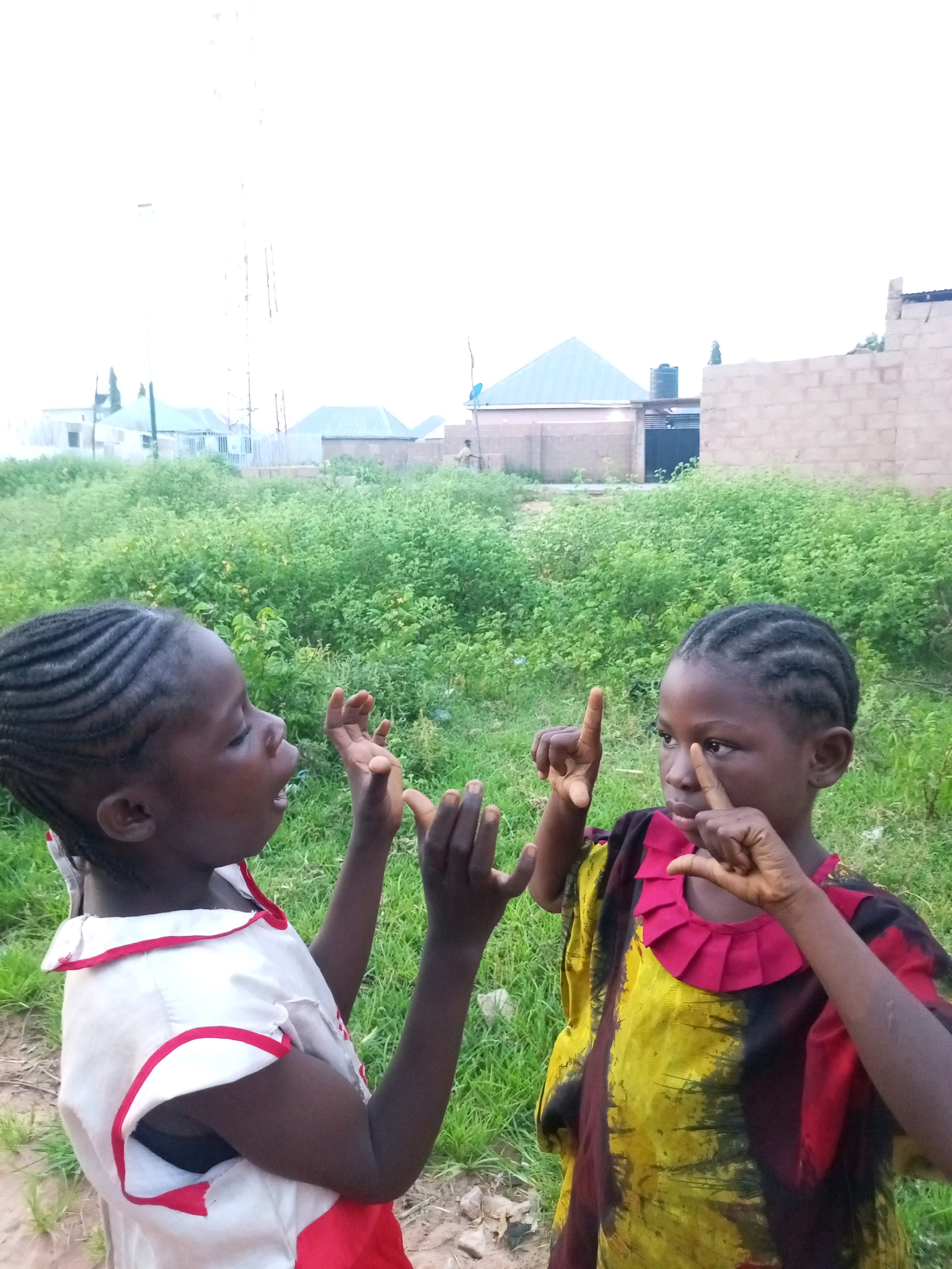 Two young girls are shown in an outdoor setting with abundant green vegetation. The girl on the left, wearing a white dress with red trim, faces left and appears to be speaking with her mouth open. Her hands are raised in front of her, with fingers splayed. The girl on the right, wearing a colorful yellow, red, and black patterned dress with a red ruffled neckline, faces forward and is looking to her left. She holds her hands up, with her index fingers extended and touching her forehead. In the background, there are buildings and a large telecommunications tower, indicating an urban or semi-urban environment. The sky is overcast, and the lighting suggests it is daytime. The scene appears to be in Tempure, Nigeria, based on the provided location information.