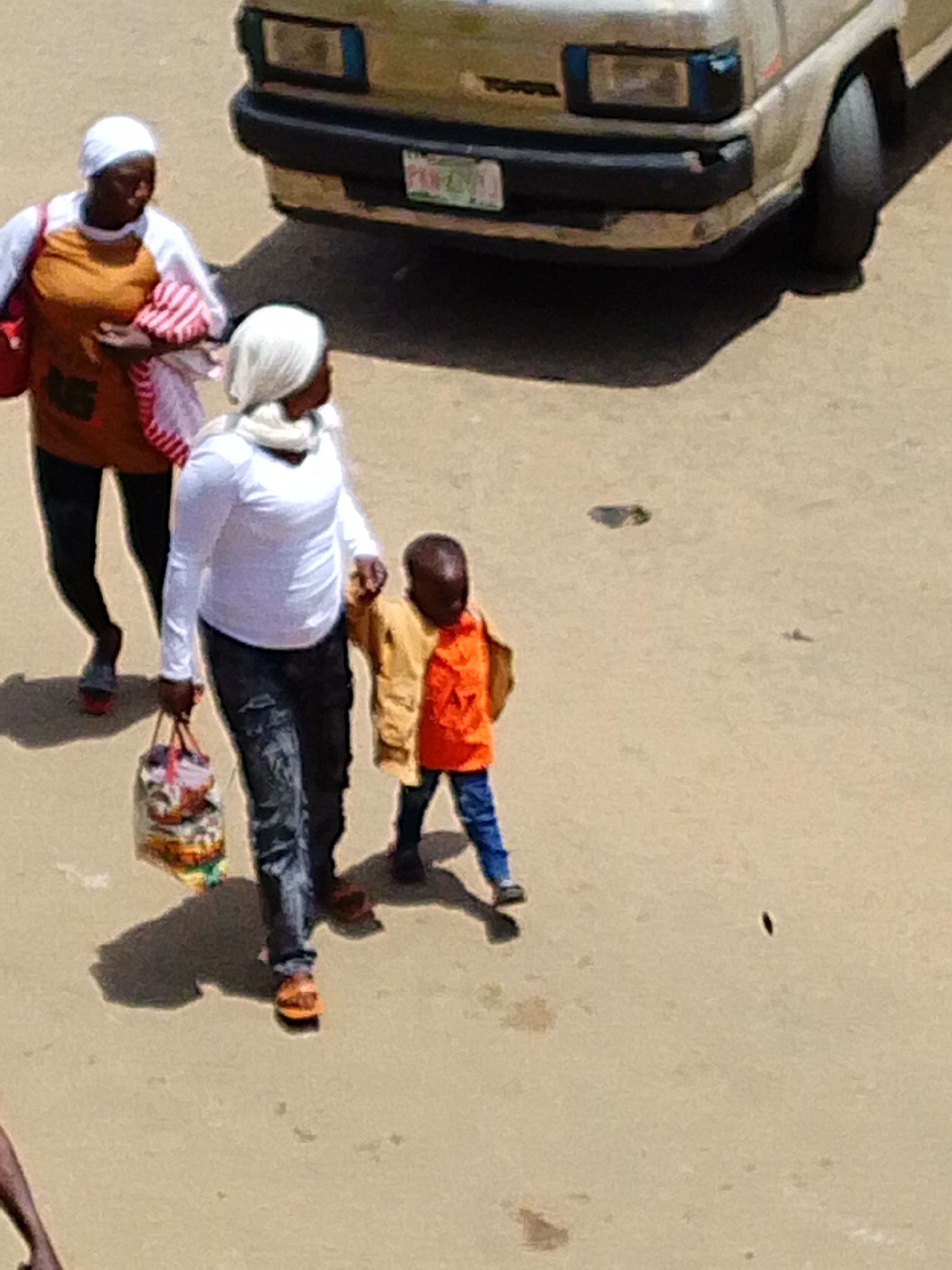 In an outdoor, presumably urban environment, three individuals are walking on a paved surface under bright sunlight, casting shadows. In the background, a light-colored Toyota van is parked. In the foreground, on the left, an adult wearing a white headscarf and an orange garment with a red and white striped cloth draped over their arm is walking. To their right, another adult, also wearing a white headscarf, is walking with a young child. This adult is dressed in a white long-sleeved shirt and dark, possibly distressed denim jeans, and is carrying a multicolored bag in their left hand. The child, on the right, is wearing a yellow jacket over an orange shirt and blue pants. The child is holding the hand of the adult in the white shirt. The ground is a sandy or dusty asphalt.