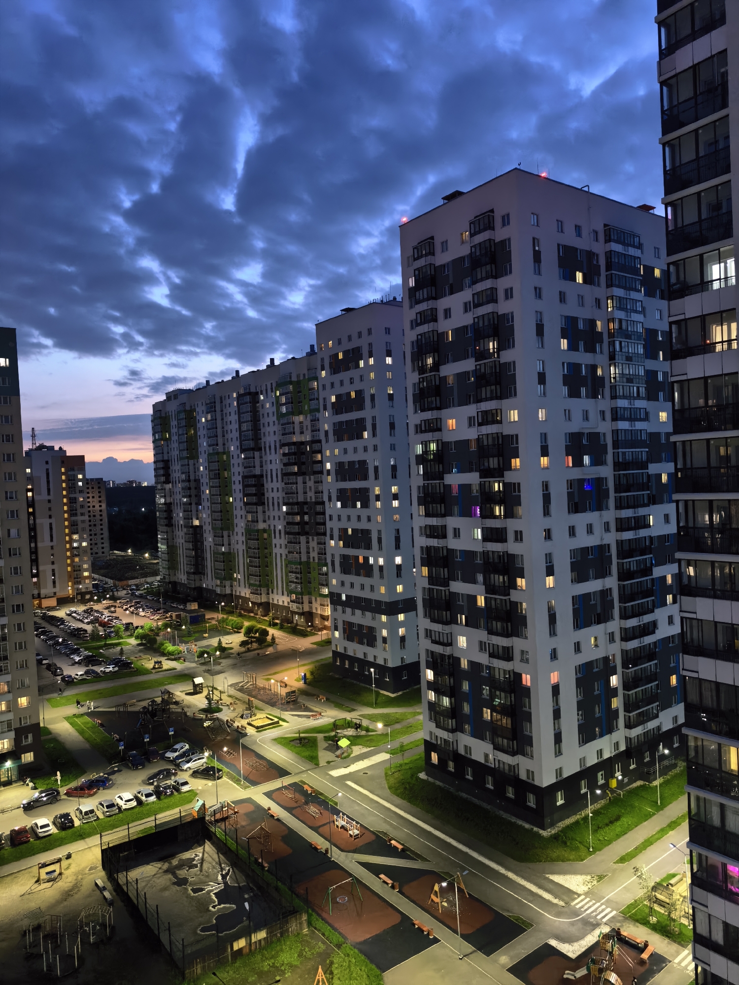 This is a high-angle, full-shot, vertical photograph of a residential complex at dusk in Unknown, Unknown. The sky is dark blue with streaks of purple and orange near the horizon, and appears to be filled with dramatic, textured clouds. The buildings are tall, modern apartment blocks with many windows, some of which are illuminated with warm, yellow light, and a few with colored lights. The architecture is characterized by clean lines and balconies.
In the foreground and midground, there are paved courtyards, playgrounds with swings and slides, and a well-lit parking lot filled with numerous cars. Green grass and trees are interspersed throughout the urban landscape. Streetlights cast pools of light on the pavement and walkways, illuminating the scene. The overall impression is of a contemporary urban environment at the end of the day. There are no visible people in the frame. The image is sharp and well-exposed, capturing the details of the architecture and the environment.