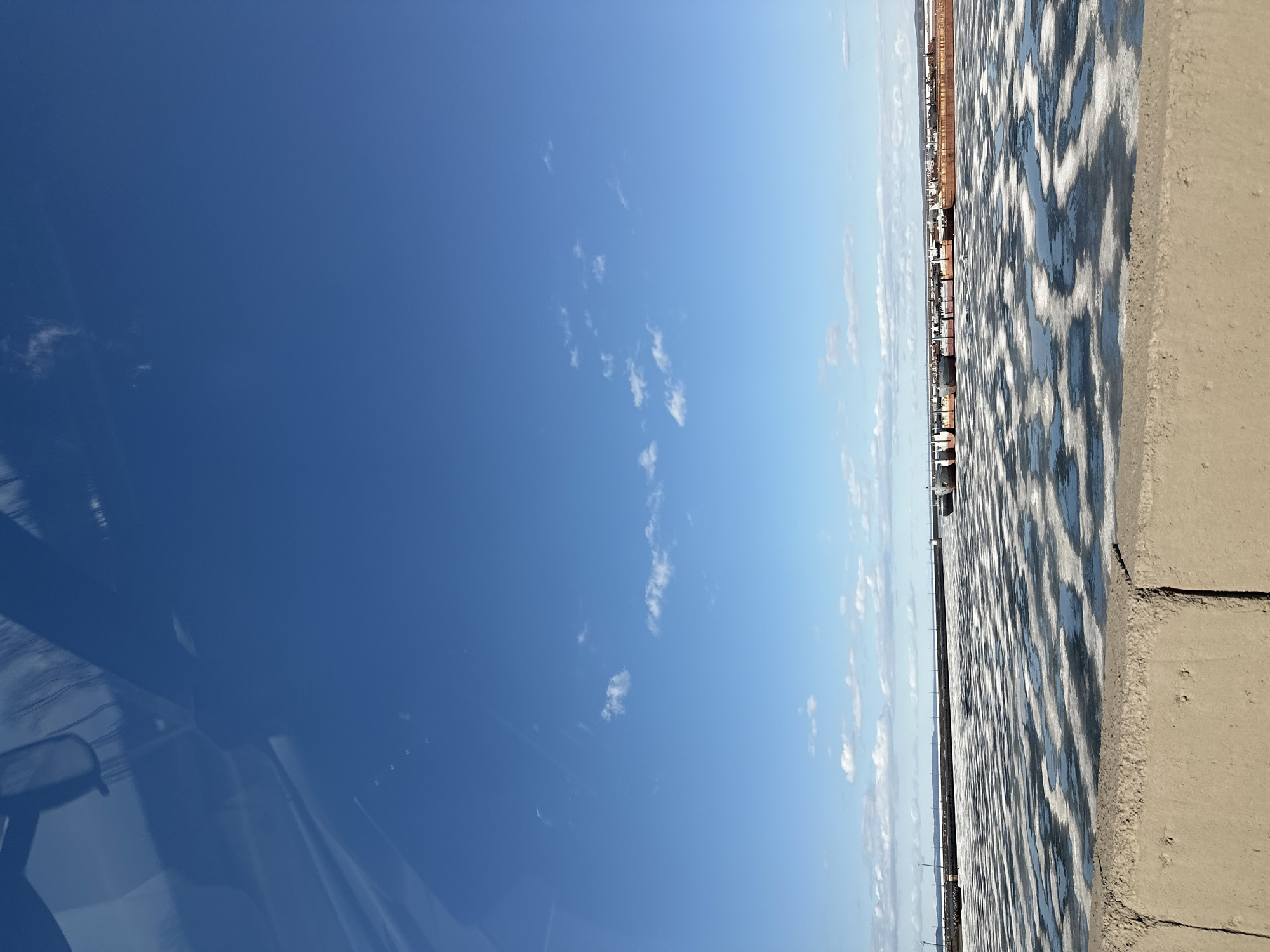The image depicts a view from a high vantage point in Chaikovsky, Russia. The lower portion of the frame is filled with a reflective surface, likely water or a similarly reflective material with alternating light and dark striations, possibly caused by ripples or ice. A concrete edge is visible on the right side of the frame, suggesting a bridge or elevated walkway. In the distance, buildings and structures line the horizon. The upper portion is dominated by a blue sky with scattered white clouds. The left side of the image includes the reflection of objects inside a car, including the rearview mirror.