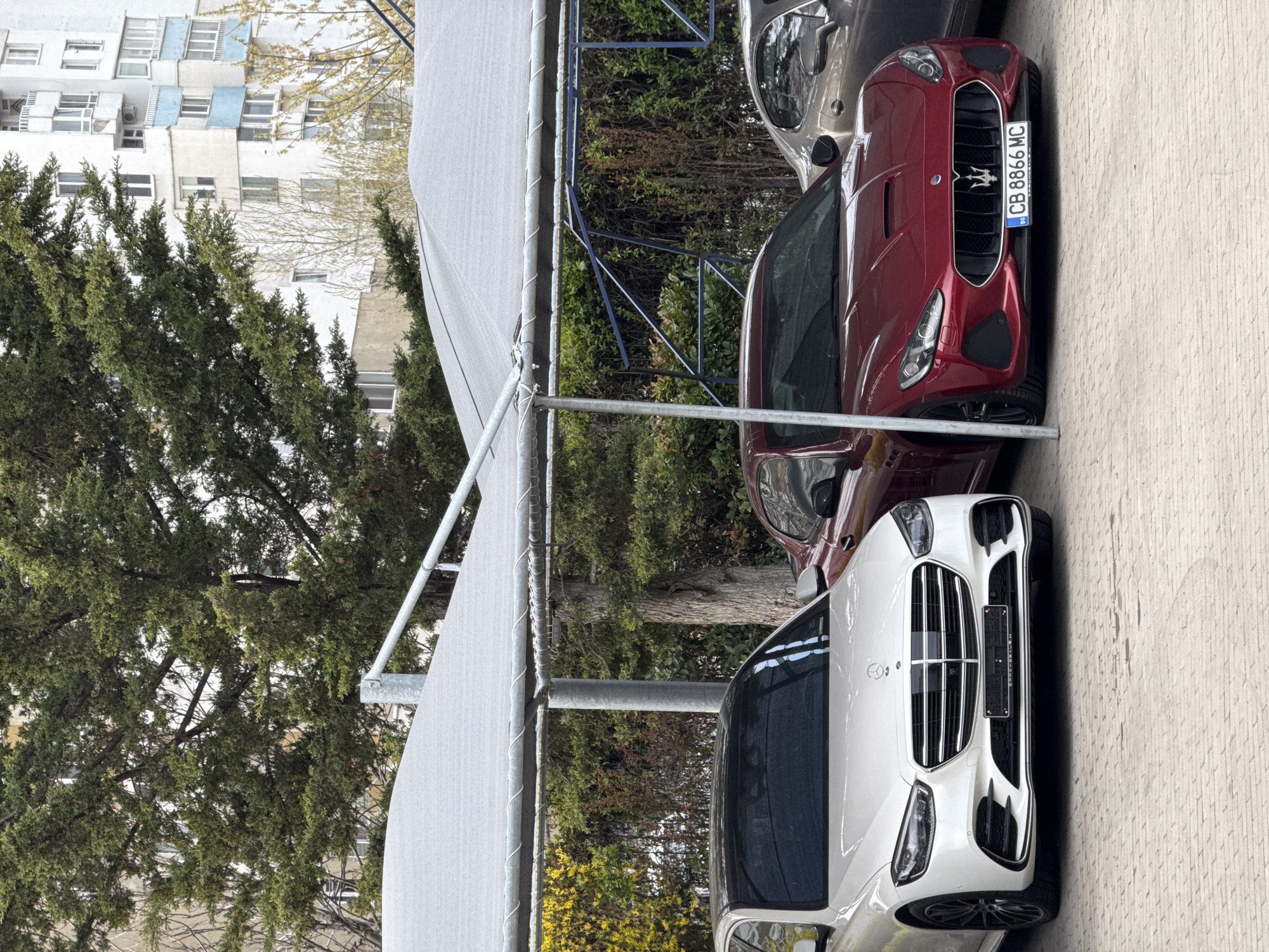 The image captures a street scene in Sofia, Bulgaria, featuring three parked luxury cars under a partial shade structure. The cars, oriented from foreground to background, are a white Mercedes-Benz SUV, a red Maserati GranTurismo, and a silver vehicle obscured further in the background. The Maserati's license plate reads "CB 8866 MC". The background includes a leafy tree and a multi-story residential building with white and blue facades. The pavement is a light, textured concrete. The photo appears to be taken during daylight hours.