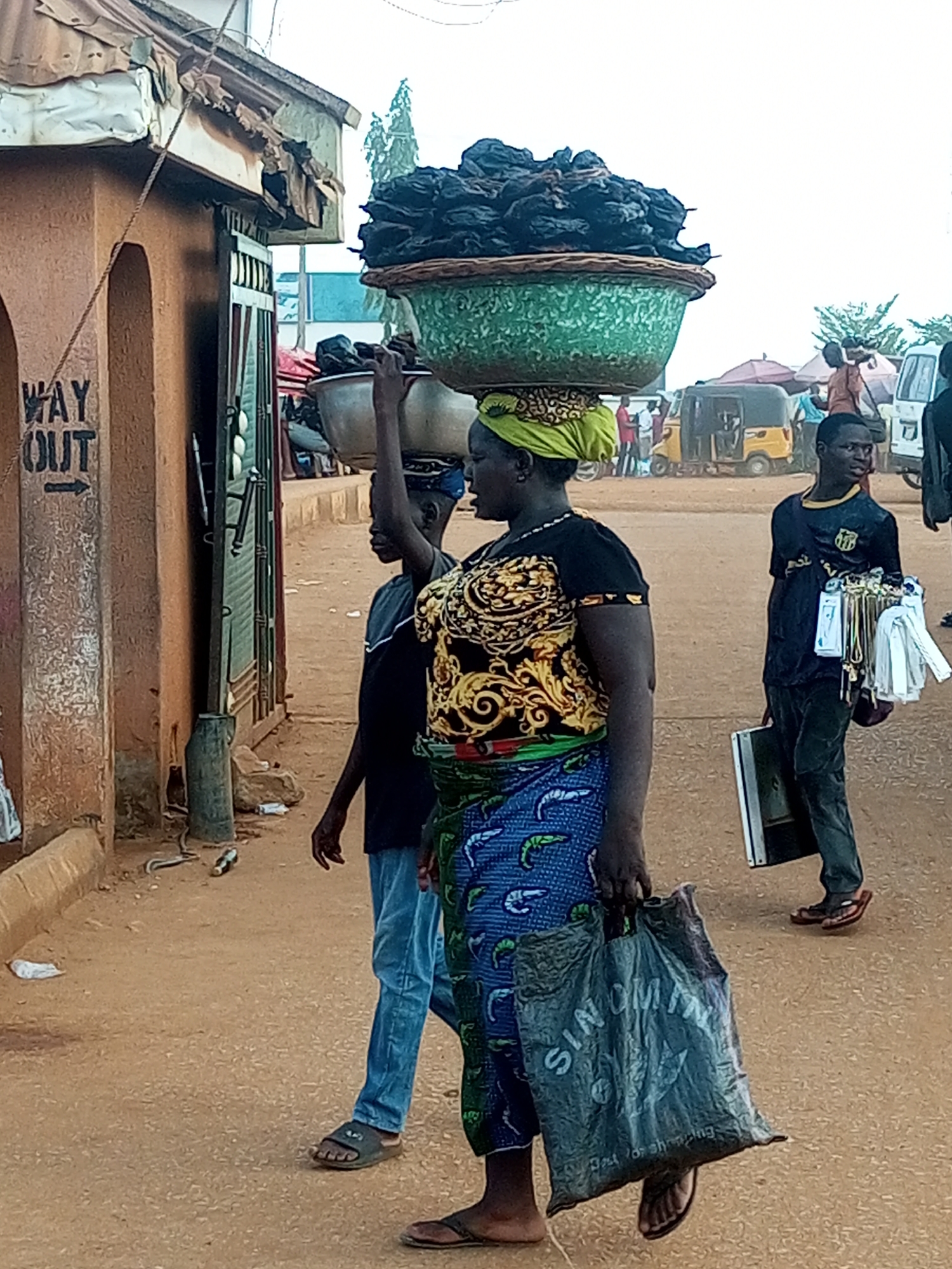 Here is a detailed description of the image:
A woman carrying a large basket filled with dark, indistinct objects on her head walks on a dirt road. She is wearing a patterned top, a blue wrap, and sandals. She is also carrying a gray plastic bag in her left hand. To her left, a younger person is also carrying something on their head, possibly a metal bowl. To her right, a person in a soccer jersey carries a briefcase and white items.
The setting appears to be a busy street in an urban area of Unknown, Unknown. In the background are vehicles, including a three-wheeled taxi, and several other people. A building with a "WAY OUT" sign is visible on the left side of the frame.