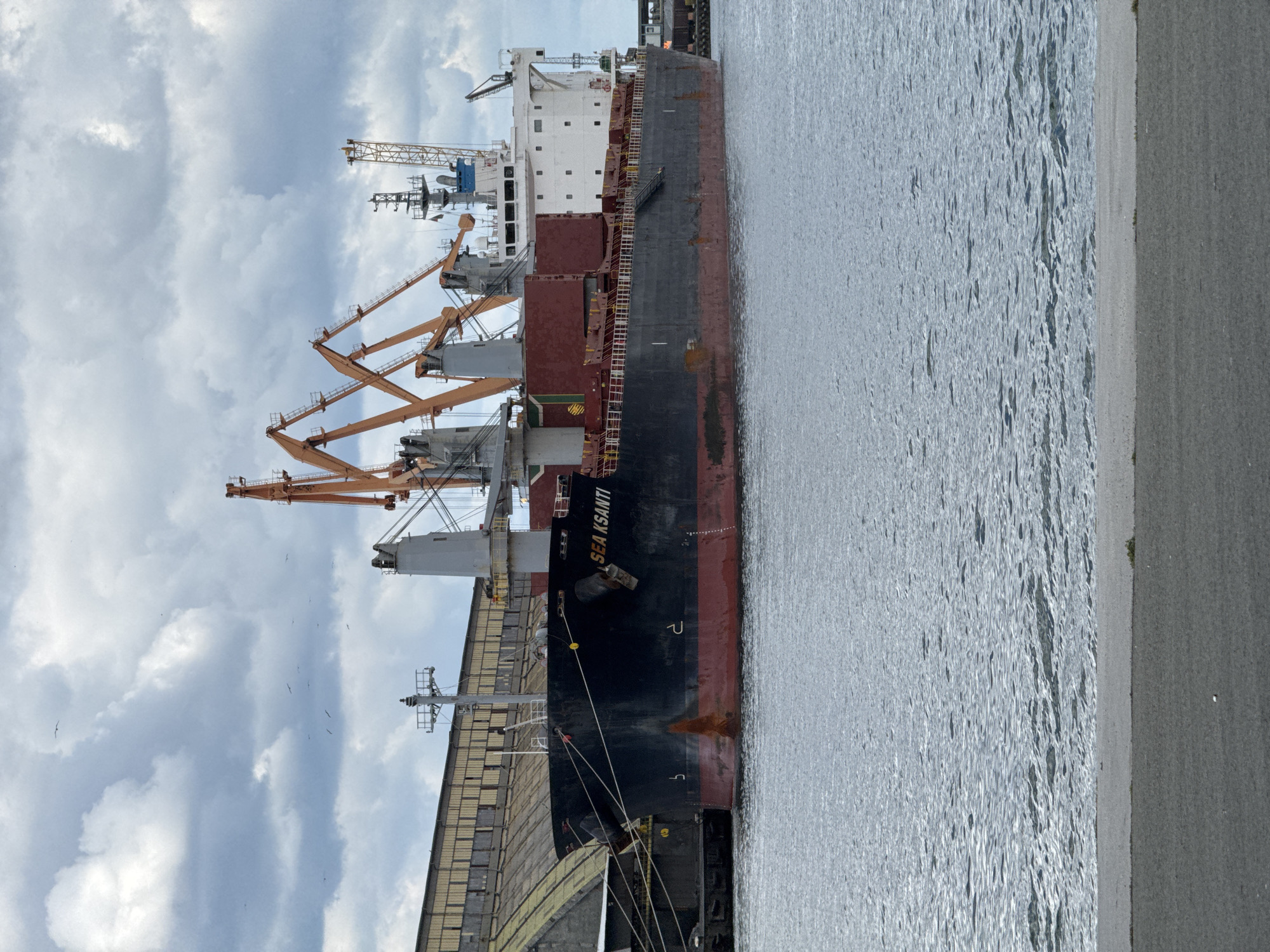 The image shows a large cargo ship named "SEA KSANTI" docked at the Port of Burgas in Burgas, Bulgaria. The ship is black and red and appears to be either loading or unloading cargo with the help of several orange cranes. The sky is cloudy. The presence of a paved area suggests that the image was taken on land.