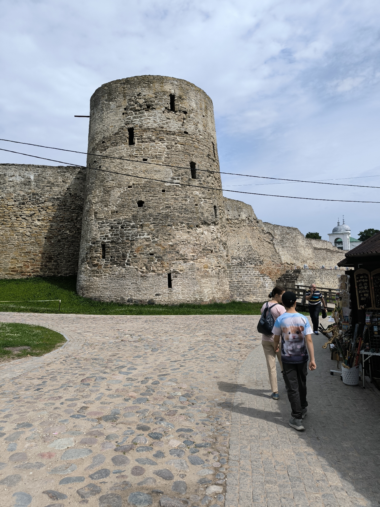 The image depicts a view of the Ivangorod Fortress, a medieval castle located in Ivangorod, Leningrad Oblast, Russia, near the border with Estonia, and not in Pechory, Russia. In the foreground, a cobblestone pathway curves to the left, bordered by a strip of green grass. Three individuals are walking along the pathway. The furthest two are a woman wearing a light-colored t-shirt and tan pants, and a young person in a black cap and a t-shirt with a hamster graphic, black pants, and grey sneakers. Ahead of them, a man in a striped shirt and dark pants walks away from the camera. To the right of the pathway, several stalls are set up, displaying various items for sale, some of which appear to be crafts or souvenirs. In the background, a section of the massive stone fortress wall dominates the scene, with a rounded tower on the left. The wall exhibits signs of age, with weathered stones and small openings. Further in the distance, a white building with a green dome is visible, possibly a church or part of the fortress complex. The sky above is partly cloudy.