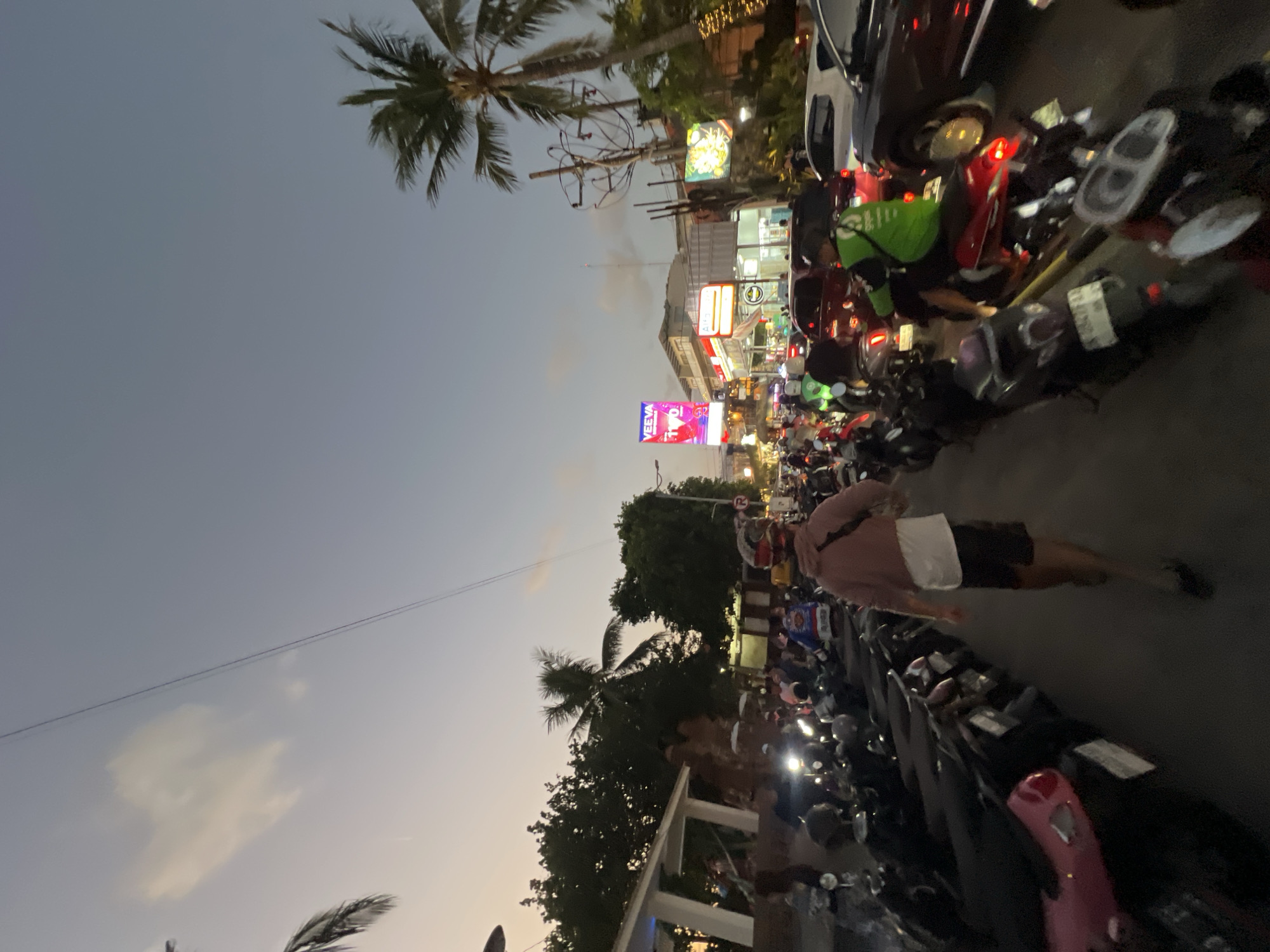 The image depicts a busy street scene in Kuta, Indonesia, at dusk. The sky is a pale blue with wispy clouds, indicating the transition from day to night. Palm trees are visible on the right side of the frame, suggesting a tropical location. The street is filled with parked and moving motorcycles, creating a sense of congestion. Several people are present, some on motorcycles and others walking. One individual in the foreground is walking away from the camera, wearing a pink hoodie and dark shorts. In the background, there are buildings with illuminated signs, including one that reads "VEEVA". Traffic appears to be heavy, with cars and more motorcycles visible. The overall atmosphere is one of a bustling urban environment during evening hours.