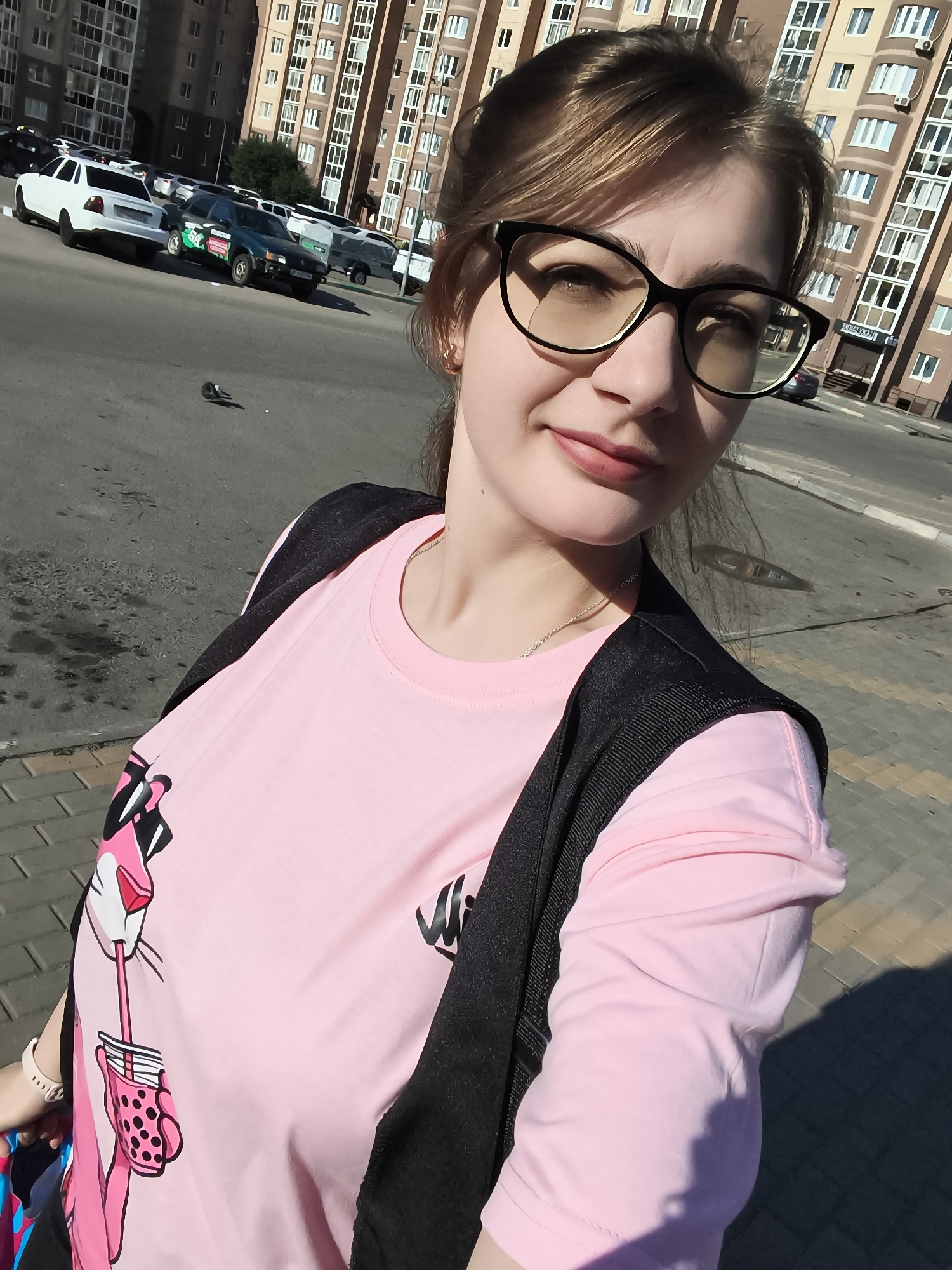 A woman wearing glasses and a pink t-shirt with a Pink Panther graphic is taking a selfie outdoors. She is standing on a paved sidewalk with a street and parked cars behind her. A tall, multi-story building with many windows is visible in the background. The woman is wearing a black vest over her t-shirt and has her hair pulled back. The lighting suggests it is daytime.