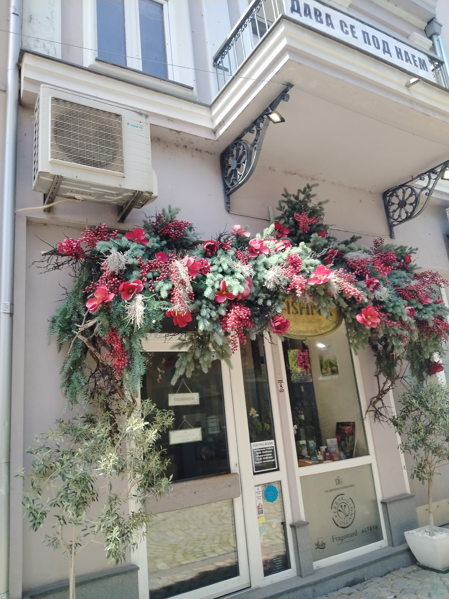 The image shows the facade of a small shop in Plovdiv, Bulgaria. The shop, "ISHMI Beauty in Nature Cosmetics & Perfume," is decorated with an elaborate floral arrangement of red flowers, berries, and evergreen branches over the entrance. A sign on the window indicates that the shop sells "Fragonard ALTEVA" products. To the left of the entrance, an air conditioning unit is mounted on the wall. Small potted trees flank the shop's entrance. Above the shop, a balcony bears a sign in Cyrillic script that appears to say "дава се под наем", which translates to "for rent." The building is painted a light gray color. The location is identifiable by the local signs and storefronts.