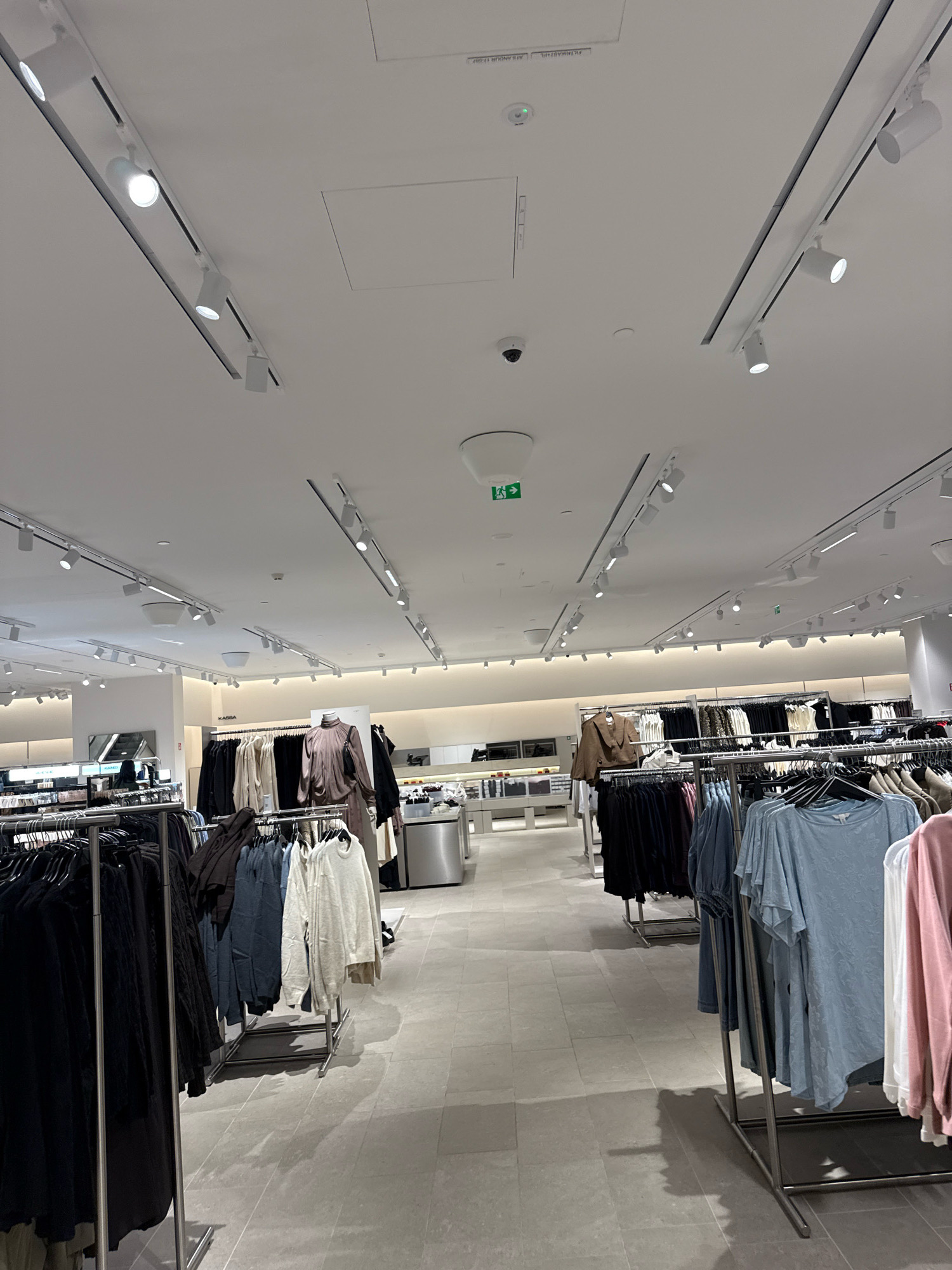 The image depicts the interior of a retail clothing store, likely a fashion boutique or department store. The scene is characterized by rows of clothing racks displaying various garments, including shirts, sweaters, and dresses, hung on hangers. The store is well-lit by a system of ceiling track lighting, with multiple spotlights illuminating the merchandise and the spacious, light-colored tiled floor. Some displays feature mannequins dressed in select outfits. In the background, there appears to be checkout counters or display tables with additional products. The overall ambiance is clean, modern, and organized, typical of a contemporary retail environment. There is no direct indication of the specific location within Tallinn, Estonia, as no exterior views or signage identifying the city are present. However, the interior design and merchandise suggest a modern retail establishment, consistent with shopping centers or high street stores found in urban centers like Tallinn.