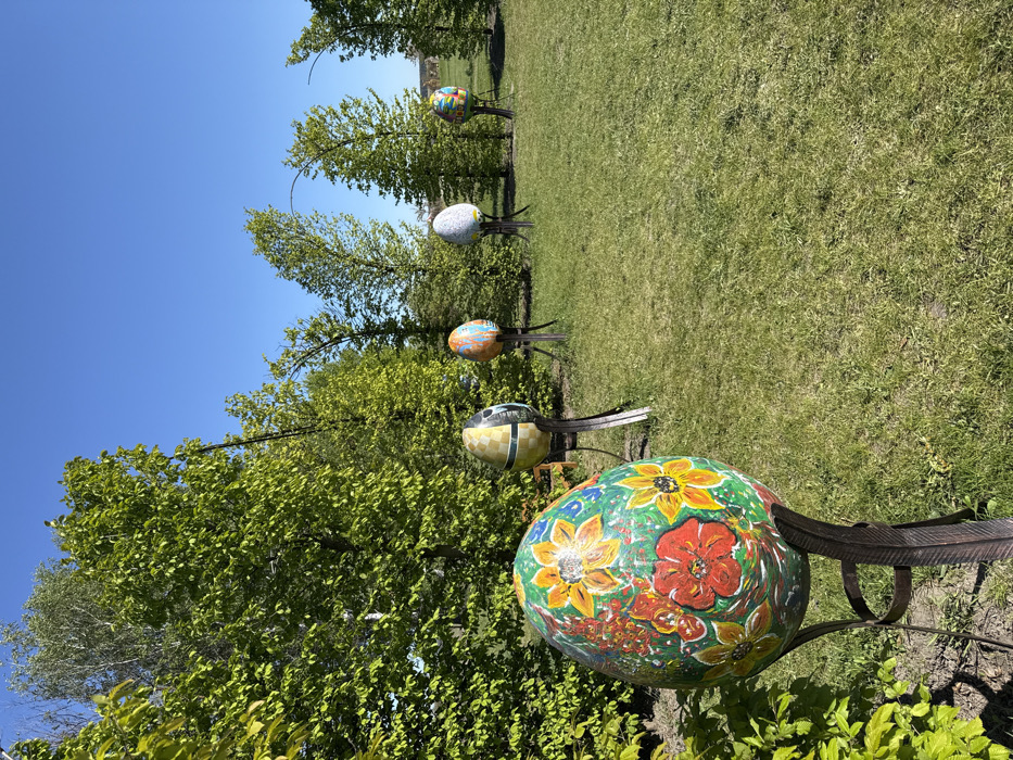 The image depicts a scene of five large, brightly colored, painted eggs on metal stands in an open grassy field. The eggs are arranged in a straight line, from left to right, on the edge of a tree line. The largest and closest egg, which is in the foreground of the image, is painted with red, yellow, and green flowers on a green background. The egg directly behind it is painted in a mosaic of yellow, white, and gray squares. The third egg, in the middle, is painted in a bright yellow and orange pattern. The fourth egg is painted mostly white with a blue and yellow design on one side and the fifth, farthest egg is red, yellow, and white with a white, blue, and red pattern on the opposite side. All of the eggs are painted with a glossy finish. The ground is covered in short, green grass with no visible weeds or bare patches. The grass is well-maintained and appears to be recently cut. The trees in the background are deciduous and appear to be in full leaf, with no signs of dead leaves or branches. The sky is a clear blue and free of clouds. There are no people in the image, but it is a scene that would be appropriate for children.
The location of the image is Kopyliv, Ukraine, which is located at latitude 50.422, longitude 29.934. This particular location appears to be in a park or recreational area. The egg installation suggests a public art display, possibly related to an event or celebration. The vibrant colors and playful designs of the eggs indicate a lighthearted and celebratory atmosphere.