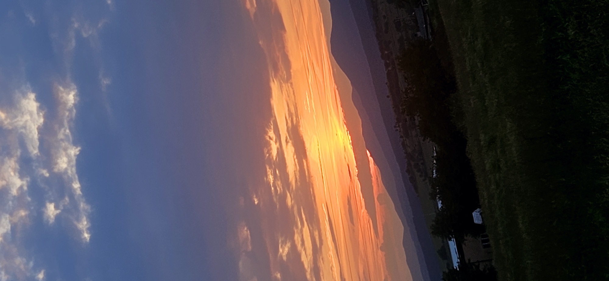 The image depicts a sunset over a mountainous landscape in Momin Prohod, Bulgaria. The sky is divided horizontally: the upper portion is a deep blue with scattered dark clouds, some exhibiting white and light-colored edges. Below this, the sky transitions into a vibrant display of orange, red, and yellow, characteristic of a sunset, with numerous elongated clouds illuminated by the setting sun. The sun itself is partially obscured by the horizon and clouds, emitting a strong golden-orange glow. Multiple layers of mountains form the midground, appearing in shades of deep blue and purple due to atmospheric haze and the receding light, silhouetted against the brightly colored sky. Below the mountain range, hilly terrain is visible with scattered dark green foliage and what appear to be small, indistinct structures or buildings. The foreground consists of a dark green, grassy field, heavily shadowed, with some faint architectural forms visible on the far left. No individuals or specific actions are discernible.