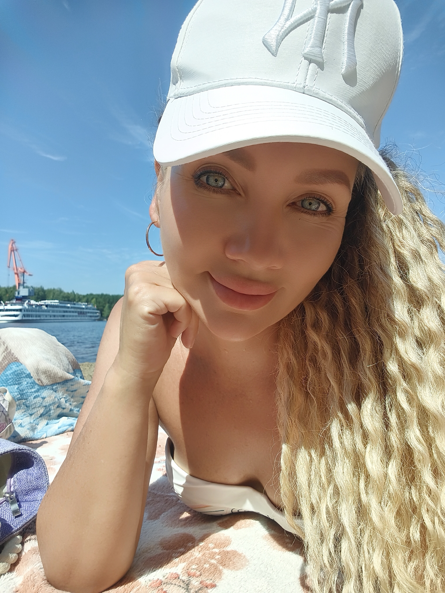 In Chaikovsky, Russia, on a sunny day, a woman with blonde curly hair lies on a patterned blanket near a body of water. She wears a white baseball cap and a white swimsuit top, with a silver hoop earring in her visible ear. Her right hand rests on her chin, and she is looking towards the camera. In the background, a large white passenger ship is docked near a red crane, with a treeline and blue sky beyond. A purple sandal is visible on the left side of the frame.