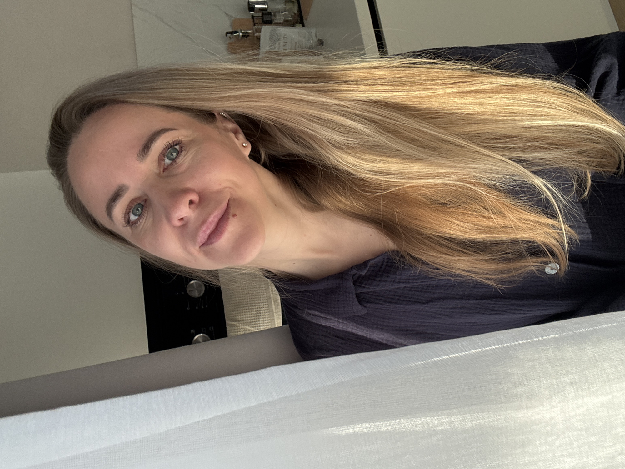 A young woman with light skin and long, flowing blonde hair is captured in a close-up, slightly upward-angled shot. Her hair, appearing to have highlights or balayage, cascades over her left shoulder and chest. She has striking blue-green eyes, neatly defined eyebrows, and a gentle, pleasant smile. A small, silver stud earring is visible in her upper left ear cartilage. She is wearing a dark blue or black textured top with visible buttons.
The woman is looking directly at the camera, suggesting a self-portrait or selfie. The background reveals a modern indoor setting, likely a kitchen, with white countertops or cabinets. On a shelf or counter behind her, glass containers are visible, with faint text "NUTS" discernible on a label on one of them. To her left, a dark appliance, possibly a stovetop or oven, with circular silver knobs, is partially visible. The lower portion of the image is dominated by a translucent white curtain or blind, suggesting a window or natural light source nearby. The overall lighting is bright and natural, indicating daytime. The scene appears to be taking place in an indoor, domestic environment in Chaikovsky, Russia.