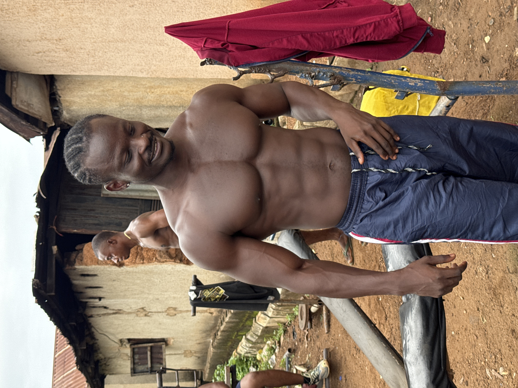 A shirtless man with defined musculature, wearing blue athletic pants, stands outdoors. The backdrop appears to be a residential area in Akwanga, Nigeria, judging by the building style. There's laundry draped over a metal rail behind him, including a red jacket and a yellow bag. Another man is seen in the background, also shirtless. The ground is dirt. The image suggests an outdoor workout setting.