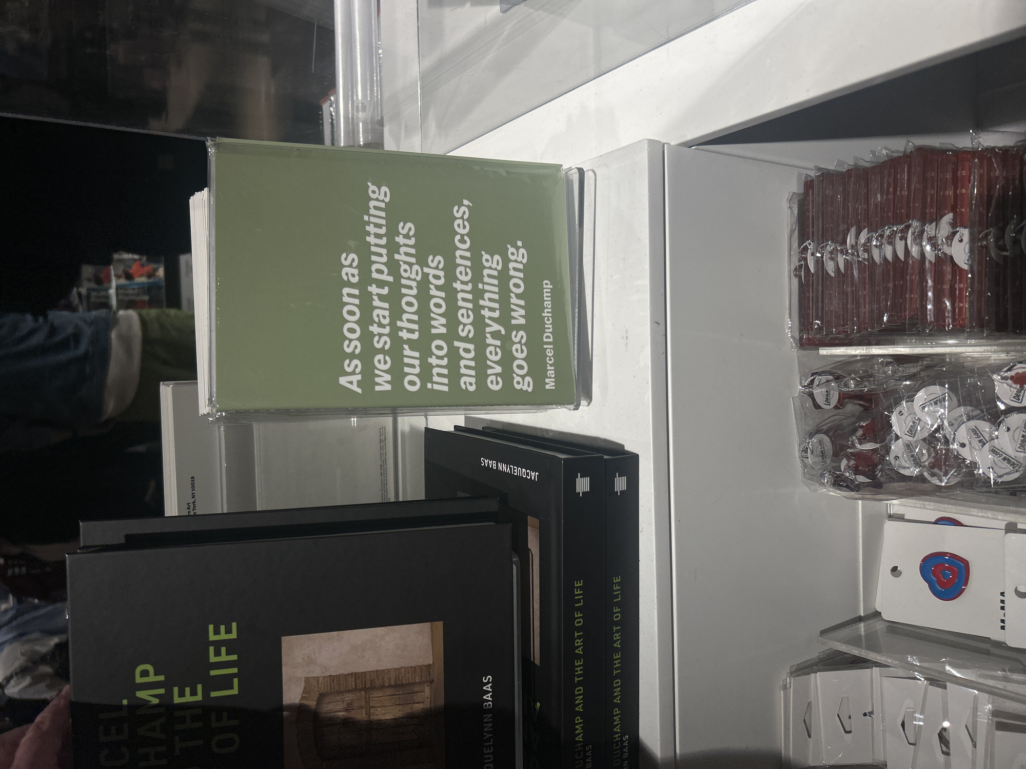 The photograph shows a display shelf, seemingly in a store, featuring a selection of books and trinkets. Two book titles are partially visible: "Marcel Duchamp the Of Life" and "Duchamp and the Art of Life" by Jacquelynn Baas. A green-covered book has the quote "As soon as we start putting our thoughts into words and sentences, everything goes wrong. Marcel Duchamp." The address "New York, NY 10019" can be discerned on a book cover. The shelf also contains small packaged items such as candies, pins, or other small accessories.
