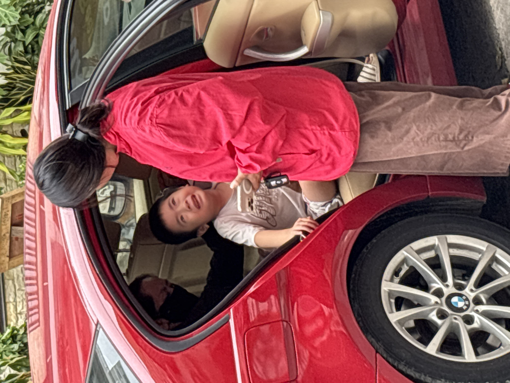 A young boy, appearing distressed and crying, is seated in the back of a red car. A woman in a bright pink jacket stands by the open car door, holding a smartphone and a keychain with a black fob. Another person wearing a black face mask is partially visible in the driver's seat. The car's interior appears to be tan leather. The scene is set outdoors, with foliage and a brick wall visible in the background, suggesting an urban environment. The presence of a BMW logo on the car's wheel indicates the car's make. The image captures a moment of emotional interaction between the woman and the crying child, with the other individual in the car as a passive observer. The setting does not provide specific visual cues to pinpoint the exact location within Nha Trang, Vietnam, beyond it being an outdoor, possibly residential or public area with a parked car.