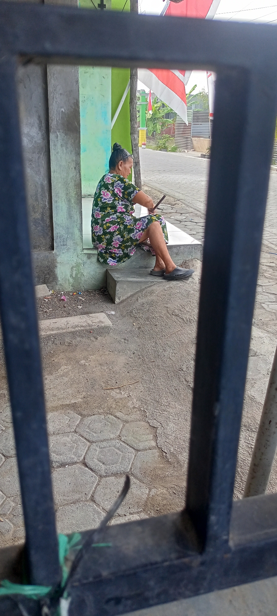 In Gempolklutuk, Indonesia, a woman with her hair tied up in a bun sits on the concrete steps of a building. She is wearing a dark green dress with a floral pattern of pink and white flowers. Her legs are crossed, and she is holding a dark object, possibly a phone or a tablet, in her hands. She is wearing dark slip-on shoes. The building behind her has a light green and grey facade. The ground in front of the steps is paved with hexagonal grey stones and concrete. In the background, Indonesian flags are visible, along with trees and a road. The scene is viewed through a dark metal grate or fence, with the bars partially obscuring the view.