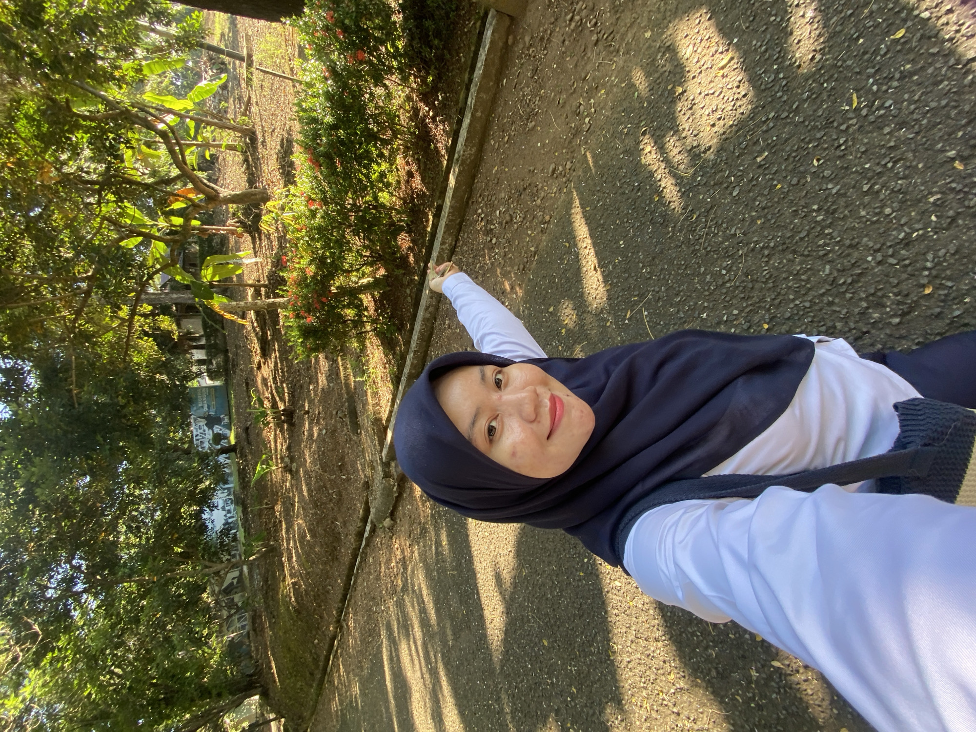 In the city of Banjarbaru, Indonesia, a woman wearing a dark blue hijab and a white long-sleeved shirt is captured in a selfie. She is standing outdoors on a paved path, with lush greenery surrounding her. The path is partially shaded by the trees, creating dappled light patterns on the ground. In the background, there are more trees, some with large leaves, and a low hedge with red flowers. A dark-colored bag is visible hanging from her shoulder. The woman is looking at the camera and smiling, with her right arm extended to the side. The overall scene suggests a pleasant day spent in a natural, outdoor setting.