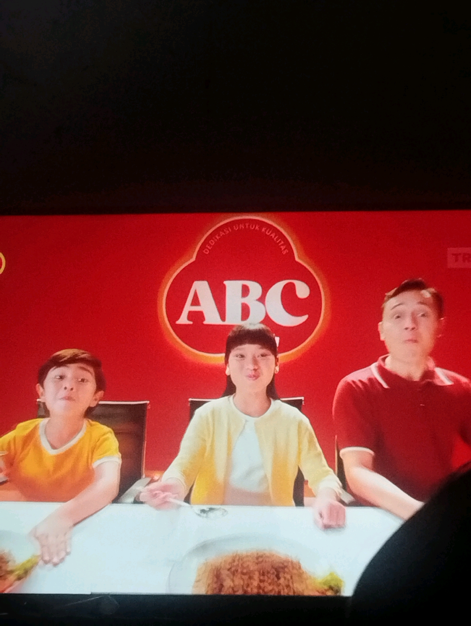In this image, three people are seated at a white table in front of a red backdrop that features a circular logo with the letters "ABC" prominently displayed. To the left, a young boy wearing a yellow t-shirt is looking towards the viewer with a curious expression. In the center, a young girl with dark hair, wearing a yellow cardigan over a white top, is looking down and appears to be holding a spoon. To the right, a man in a red polo shirt is looking towards the viewer with a surprised or shocked expression. The table in front of them has white plates with food, appearing to be rice dishes, and the overall setting suggests a mealtime advertisement or scene. This scene is likely from an advertisement filmed in South Jakarta, Indonesia, given the prominence of the "ABC" brand, a well-known Indonesian food and beverage company.