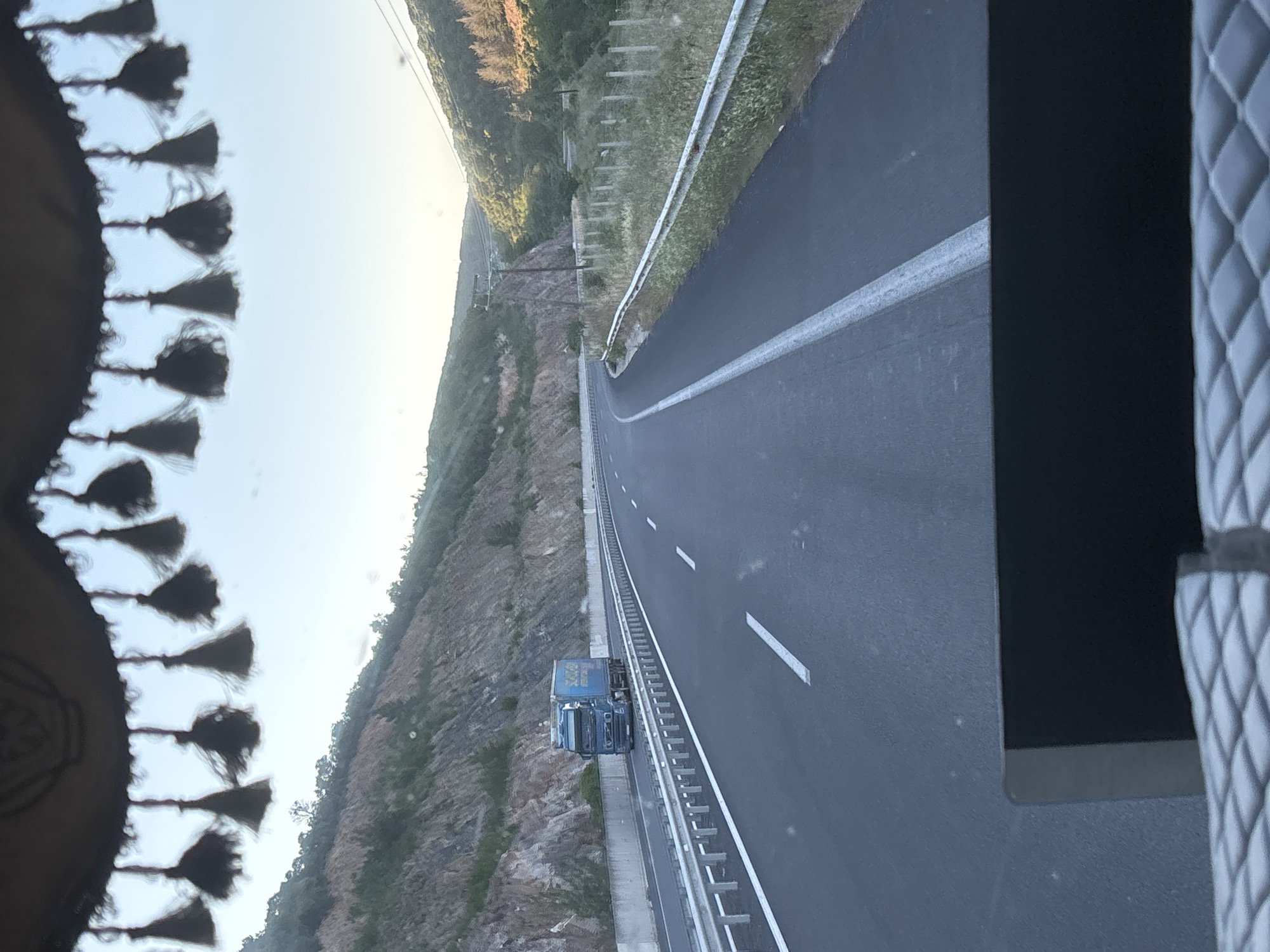 The image depicts a view from inside a vehicle looking out at a highway. A blue semi-trailer truck is traveling on the highway in the left lane. The truck has a yellow trailer with blue lettering. The highway is flanked by a concrete barrier and a metal guardrail on the left, and a grassy area with a fence on the right. The landscape beyond the highway consists of a steep, rocky hillside on the left and rolling, tree-covered hills on the right. The sky is clear and bright, suggesting it is daytime. The scene is set in or near Avra, Greece.