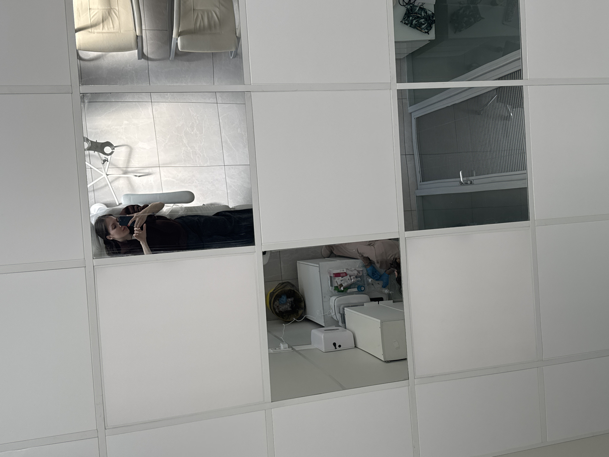 The image depicts a grid of square panels, some of which are mirrored, reflecting parts of a room. One reflection shows a person lying down, looking at a mobile phone. Another mirror reflects a window with closed blinds. A third reflected section shows a room corner with medical equipment, including a mini-refrigerator, a white box and a pet carrier. The location is in Moscow, Russia.