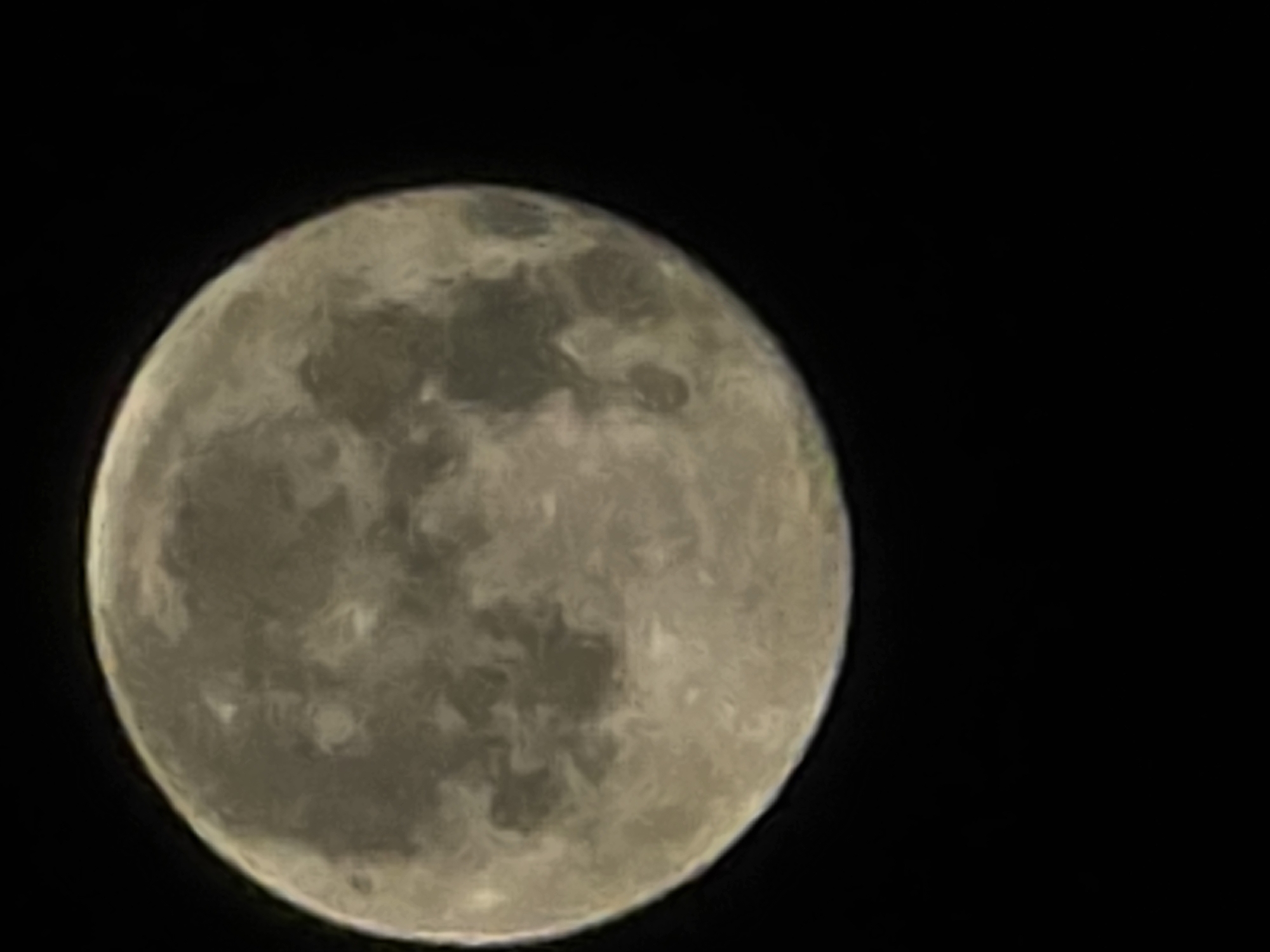 A close-up, slightly blurry photograph shows a full moon against a black sky. The moon is illuminated from the side, casting shadows that create variations in its surface texture. Areas of lighter and darker grey are visible, suggesting craters and maria. The bottom half of the image is completely black, indicating the absence of any surrounding landscape or city lights. There are no people, objects, or discernible actions taking place. The scene is purely celestial. No specific location in Yambol, Bulgaria can be identified from this image.