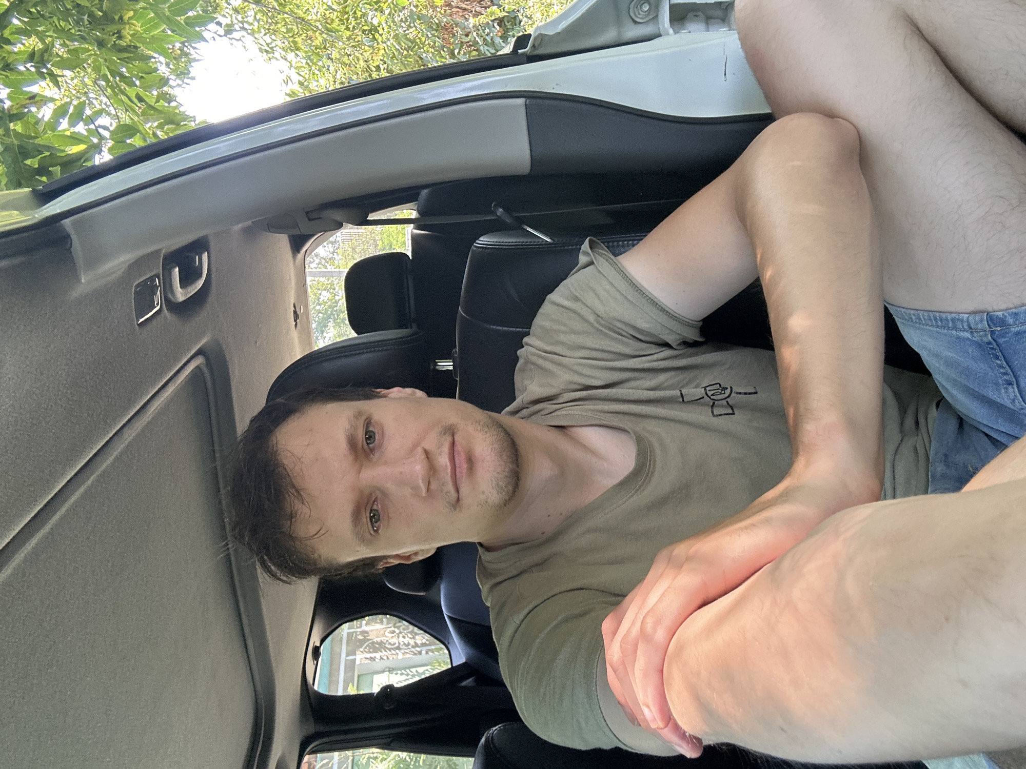 The image shows a young man sitting inside a car, likely in the city of Makiv, Ukraine. He is wearing a light green t-shirt and denim shorts. The man is looking directly at the camera, with his arms crossed and resting on his leg. Sunlight is visible through the car windows, casting shadows on his arms and legs. The interior of the car is visible, including the seats and the sunroof. Outside the car, trees and foliage can be seen, suggesting a natural or suburban setting. The overall atmosphere is casual and relaxed.