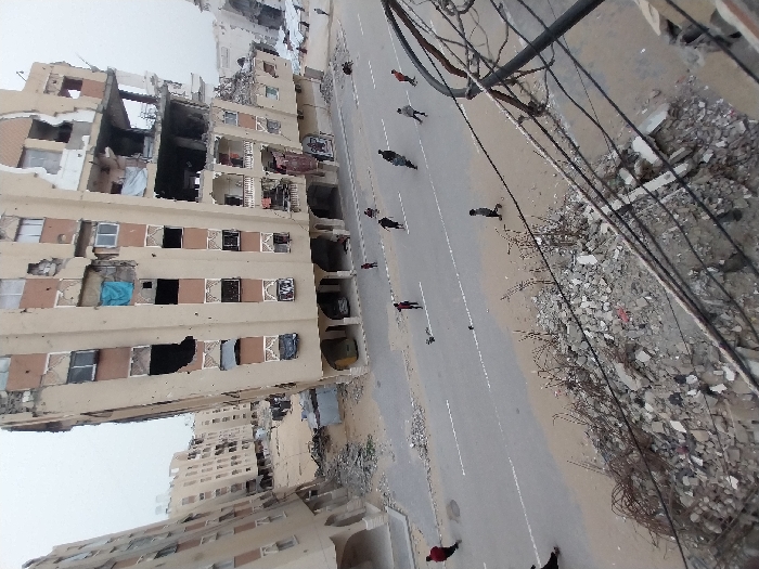 A desolate street stretches between damaged buildings. The buildings display extensive destruction, with gaping holes and collapsed sections. Individuals walk along the roadway, seemingly navigating the aftermath. Debris and rubble litter the ground, highlighting the devastation of a conflict zone. Overhead, power lines and wires form a tangled web, adding to the scene's chaotic atmosphere. The image conveys a sense of desolation and loss amidst the ruins.