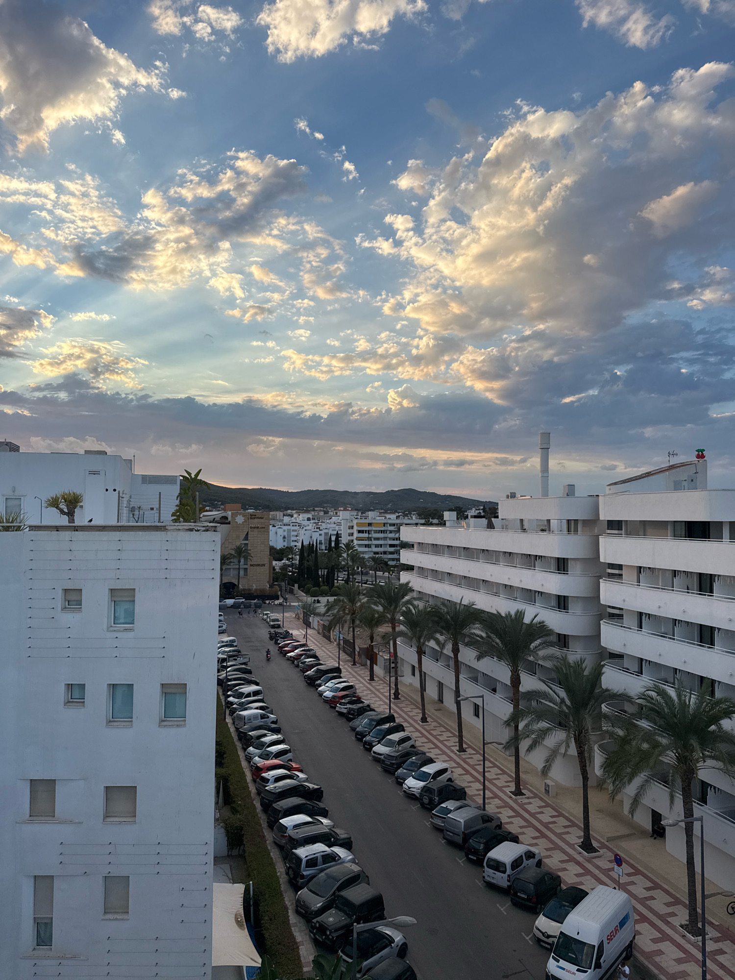 The image depicts a street scene in Ibiza, Spain, with a view of buildings and parked cars under a dramatic sky. The foreground features the side of a white building with several windows. To the right, a street is lined with parked cars, including vans, SUVs, and sedans, running parallel to a sidewalk with palm trees. Beyond the street, more white buildings with balconies and archways are visible, some with signage indicating businesses. In the background, a town with buildings and hills rises, and the sky is filled with dynamic clouds, with rays of sunlight breaking through. The overall impression is an urban coastal environment during what appears to be late afternoon or early evening.