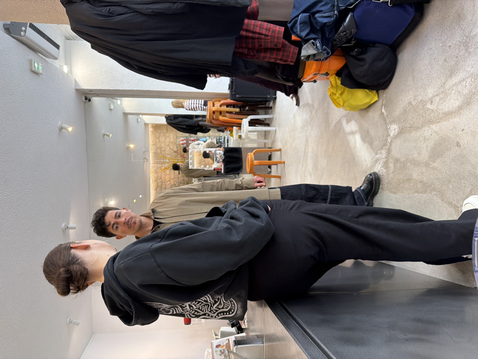 The image depicts an indoor scene within a commercial or studio space in Paris, France. This is indicated by the French "SORTIE DE SECOURS" sign visible on the ceiling.
In the foreground, a female subject with dark hair styled in a bun stands with her back to the camera, leaning against a dark, possibly metallic, counter. She wears a black oversized hoodie with a white graphic design on the back and black wide-leg trousers, with white sneakers visible at her feet. Directly facing her, in the mid-ground, stands a male subject with light skin, dark hair, and a mustache. He wears a light tan or khaki long-sleeve collared shirt over a darker undershirt, dark wide-leg jeans with visible white stitching, and black shoes. His gaze is directed towards the viewer.
The environment features a light-colored, textured concrete floor. The walls are predominantly white, with a section of exposed light-colored stone wall visible in the background. Various objects are distributed throughout the space: on the right side of the frame, several dark coats hang from a rack, beneath which are stacked numerous bags in yellow, orange, blue, and black, alongside a dark wheeled suitcase. Several orange and white stools are scattered in the mid-to-background. A large mirror on the back wall reflects parts of the room, showing another individual with a striped shirt and another near a lighted vanity mirror. Ceiling fixtures include recessed spotlights, an air vent, and the aforementioned emergency exit sign.