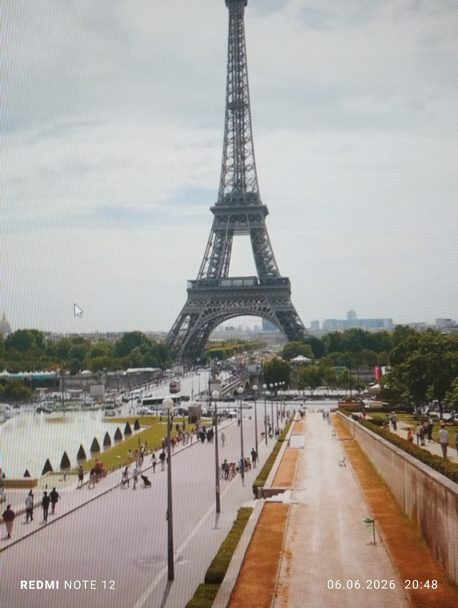 This image is a digital screenshot depicting a wide, elevated view of the Eiffel Tower in Paris, France, observed from a vantage point likely near the Trocadéro. The entire image exhibits prominent vertical scan lines, indicative of a photograph taken of a digital display, and a white mouse cursor is visible in the upper-left quadrant.
In the prominent background, the Eiffel Tower stands centrally under an overcast, light grey sky. Below the tower, the River Seine is visible, with several boats and barges docked along its banks, and a bridge crossing it.
The midground features a bustling promenade with numerous pedestrians walking in various directions. This paved area is lined with uniformly spaced lampposts and has landscaped sections with green grass, small trees, and conical shrubs. Beyond the river, more urban structures and lush green areas are discernible.
The immediate foreground reveals a lower, unpopulated pathway with earth-toned surfaces flanked by low, green hedges.
Overlay text is present in the bottom corners: "REDMI NOTE 12" appears in the lower-left, indicating the original image capture device, and "06.06.2026 20:48" is displayed in the lower-right, denoting the date and time. The scene is illuminated by ambient daylight, suggesting late afternoon or early evening.