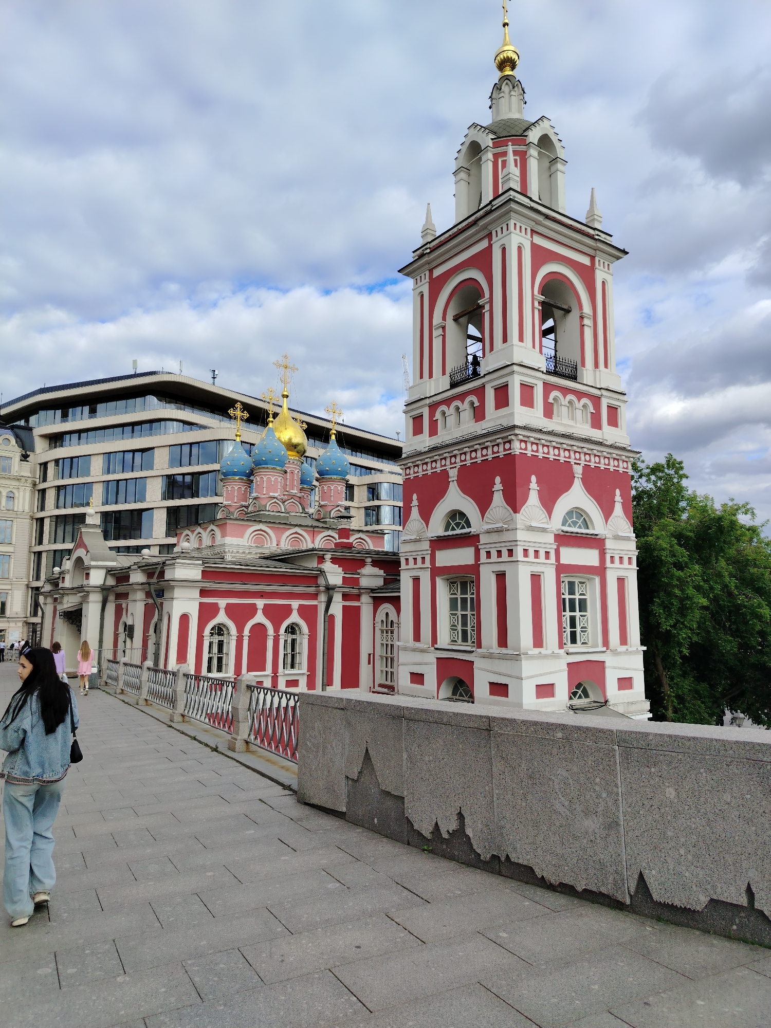 In an outdoor, urban setting, a red and white bell tower with a golden dome and cross stands prominently to the right. Adjacent to it is a red and white church with multiple blue domes topped with golden crosses. A modern, multi-story building with a glass facade is visible behind the church and bell tower. In the foreground, a woman with long dark hair, wearing a denim jacket and light-colored pants, walks away from the camera on a paved pathway. Other individuals are seen in the middle ground walking along a railing in front of the church. The sky is overcast with scattered clouds. The scene appears to be a public space with architectural elements of both historical and contemporary design.