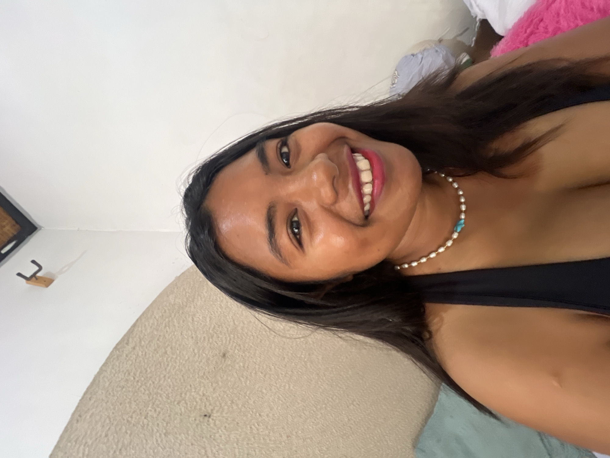 A young woman with dark hair and brown skin is captured in a close-up, smiling selfie indoors. She is wearing a black tank top and a pearl necklace with a turquoise pendant. The background includes a textured, curved wall and a white wall with a hook. The lighting appears bright, highlighting the woman's face and skin. The image is a simple, informal portrait of the woman.
