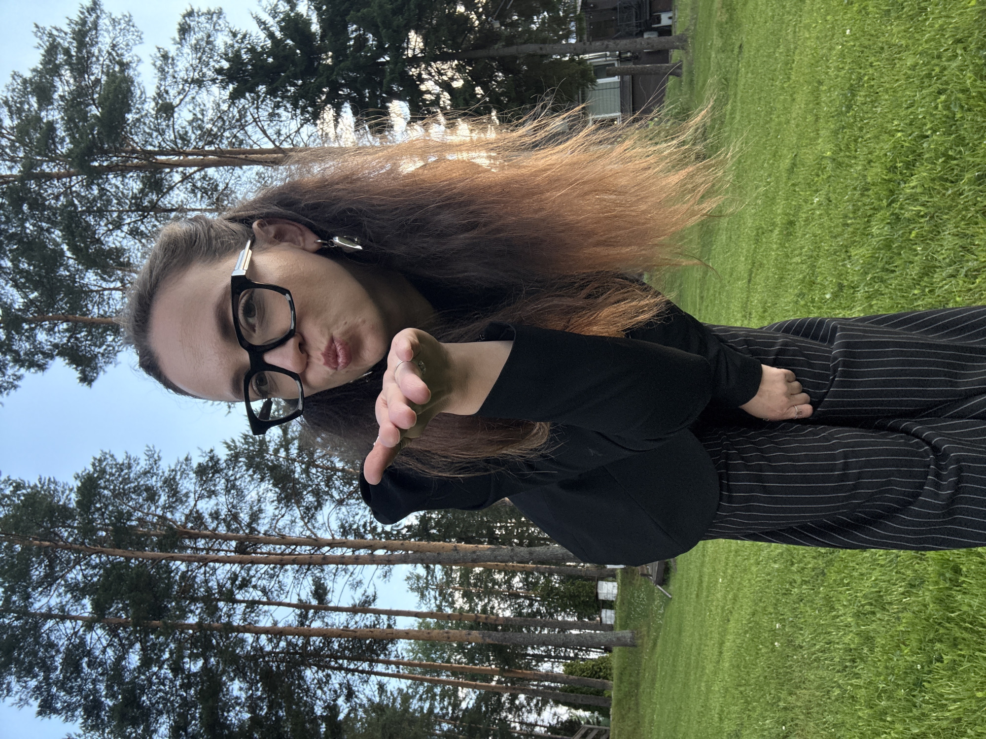A young woman with long, wavy auburn hair, wearing glasses and a black outfit with pinstripe pants, blows a kiss towards the camera. She is outdoors, with a backdrop of tall pine trees and a lush green lawn. The sky is visible above the trees, suggesting it is daytime. The scene appears to be a park or a yard. The woman's expression is playful and affectionate as she makes the gesture. There is no visible text in the image.