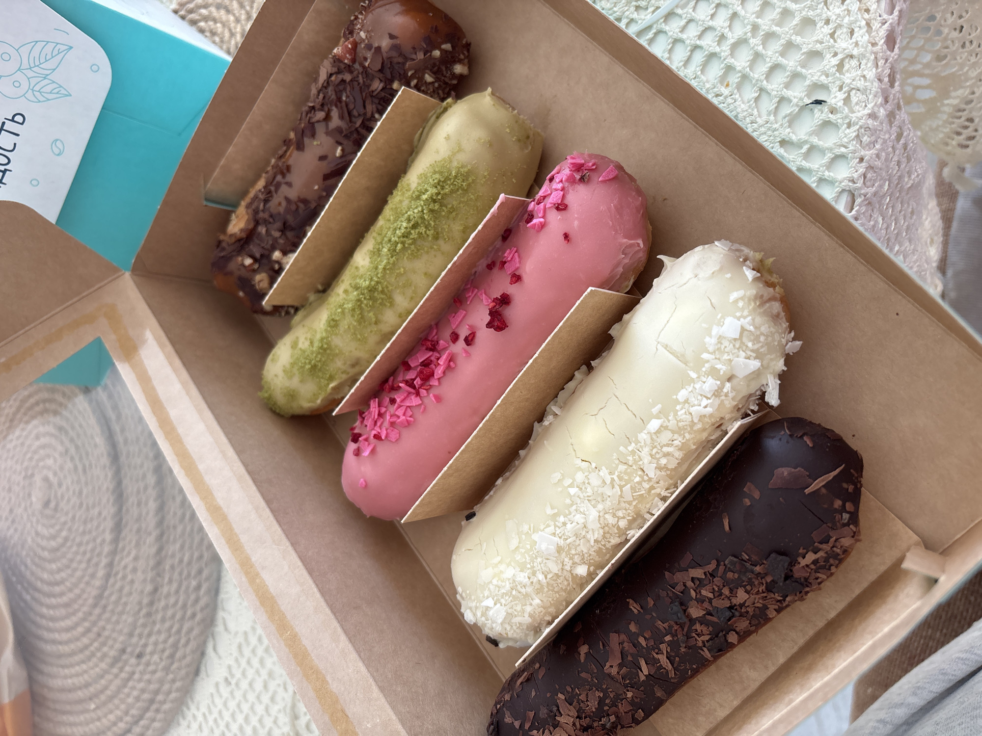 A box containing six eclairs is photographed from an overhead perspective. The box, light brown with a transparent window on the side, showcases eclairs with different colored icings and toppings. These include brown with chocolate shavings, green with green sprinkles, pink with pink sprinkles, white with coconut flakes, and dark brown with chocolate shavings. The box is set on a light surface. Part of a light blue box with some inscriptions in the Russian language is visible on the left side of the frame. The photograph was taken in Krasnoyarsk, Russia.