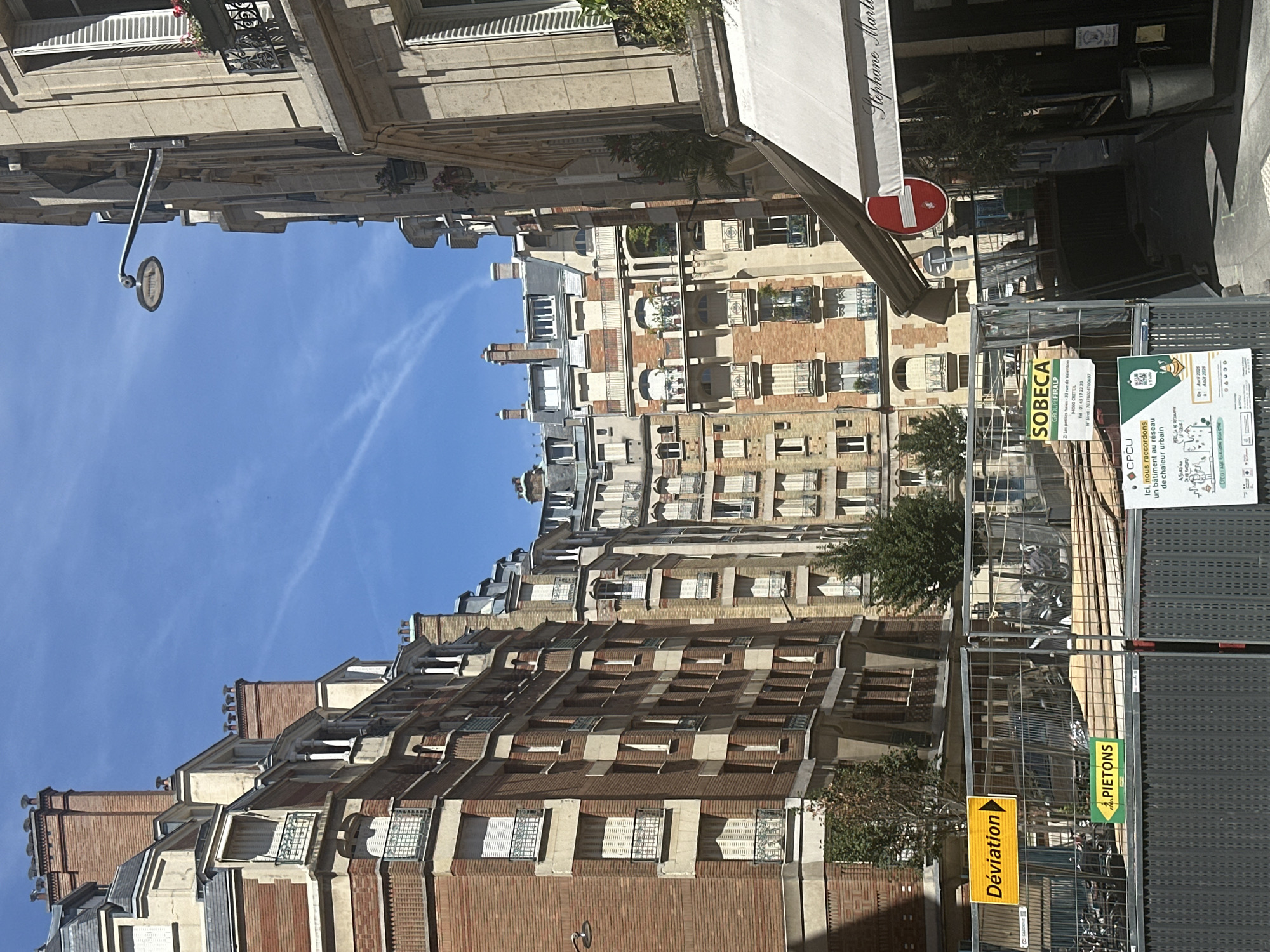 This is a street-level, eye-level, medium shot of a city street in Paris, France, on a sunny day with a clear blue sky and a few wispy clouds. The scene is dominated by tall, multi-story buildings with brick and stone facades, characteristic of Parisian architecture, featuring numerous windows and balconies.
In the foreground, there is a construction site enclosed by metal fencing. Two signs are visible on the fencing: a yellow rectangular sign with the word "Déviation" (detour) and an arrow pointing to the right, and a smaller green rectangular sign with a pedestrian icon and the word "PIETONS" (pedestrians). There are also stacks of wooden planks and construction materials behind the fence. Another sign further down the fence displays "SOBECA GROUPE FIPALP" with an address and phone number, along with construction information.
A white awning is extended from a building on the right, with the name "Stephane Marti" partially visible on it. A red and white circular "no entry" sign is hanging from the building near the awning. A street lamp with a circular fixture is mounted on the side of a building.
Several motorcycles are parked behind the construction fencing. A small tree is planted in a metallic pot near the awning. The overall impression is a busy urban environment, possibly undergoing some renovation or construction work, with a mix of residential and commercial elements.