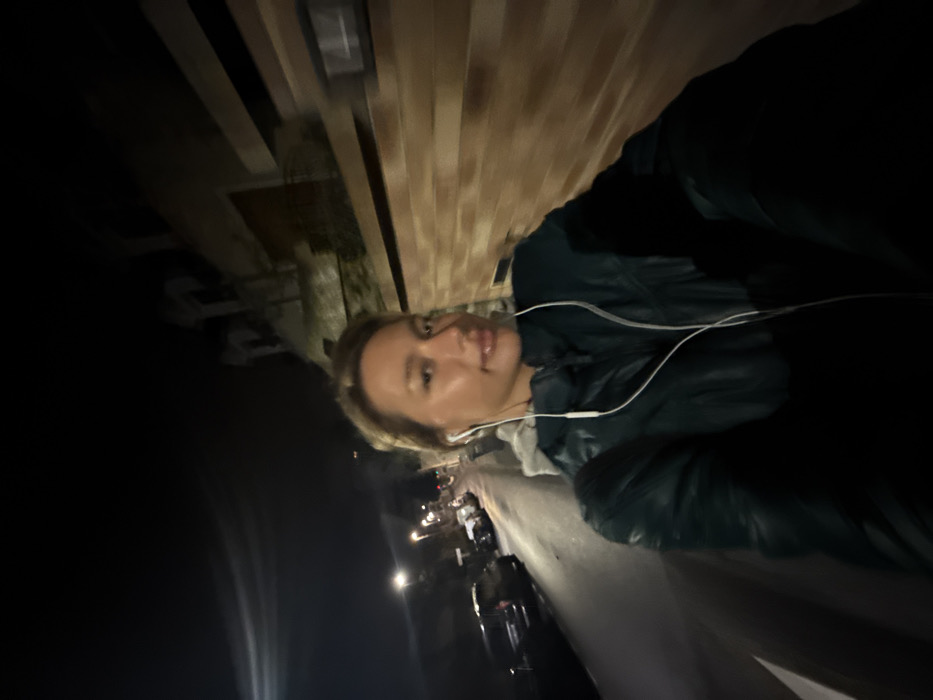 A woman with short blonde hair wears wired headphones. She's wearing a dark jacket, possibly leather, over a grey hooded top. She's on a street at night, with buildings visible behind her, some with wood siding. Cars are parked along the street. The lighting is dim, and the photo is taken at an angle.