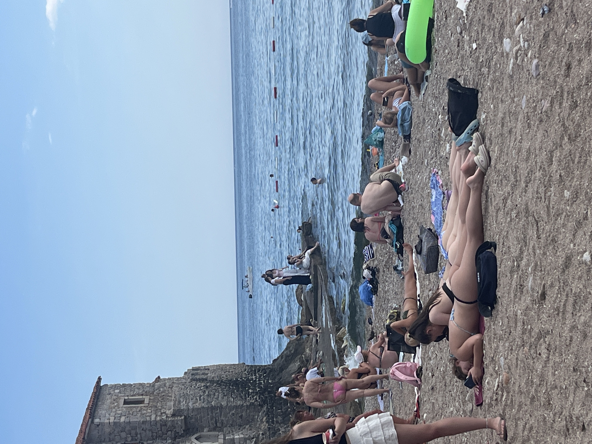 The image depicts a crowded beach in Budva, Montenegro, with a stone building on the left side and the Adriatic Sea in the background. Numerous people are sunbathing, swimming, and relaxing on the pebble beach. Some individuals are lying on towels, while others are wading in the shallow water. A boat is visible in the distance on the sea, and buoys mark a swimming area. The scene is set under a clear blue sky, suggesting a sunny day at the coast.