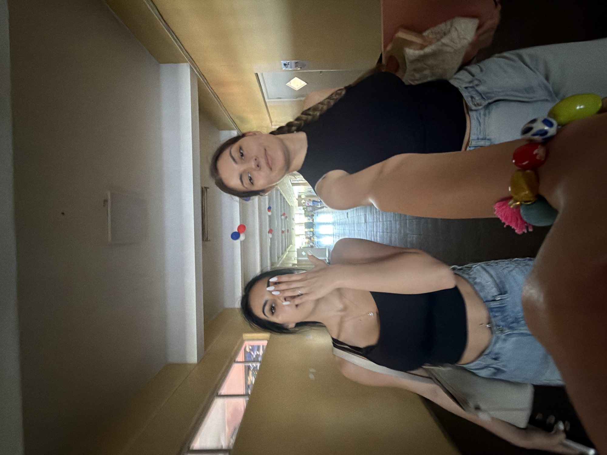 Two young women are captured in what appears to be a selfie or casual photo taken indoors in Burgas, Bulgaria. The image is rotated, showing them aligned vertically, but they are likely standing in a hallway.
The woman closer to the camera, on the left of the frame in the original orientation, has dark hair styled in a braid and is looking directly at the camera with a slight smile. She is wearing a black tank top and light-wash denim jeans. A colorful bracelet with various beads (red, yellow, blue, green) and a pink tassel is visible on her right wrist.
The second woman, further back and to the right, also has dark hair. She is partially covering her face with her hand, revealing white nail polish. She is wearing a black crop top, light-wash denim jeans, and has a belly button piercing visible. A simple necklace is also visible.
They are in a long, well-lit hallway with light brown/beige walls and a darker brown ceiling. Windows line one side of the hallway, allowing natural light to enter. Small red, white, and blue decorative items (possibly paper lanterns or balloons) are hanging from the ceiling in the distance, suggesting a festive or themed event. Other people are faintly visible further down the hallway. A number "308" is barely visible on a door or frame in the background. The overall mood appears casual and playful.