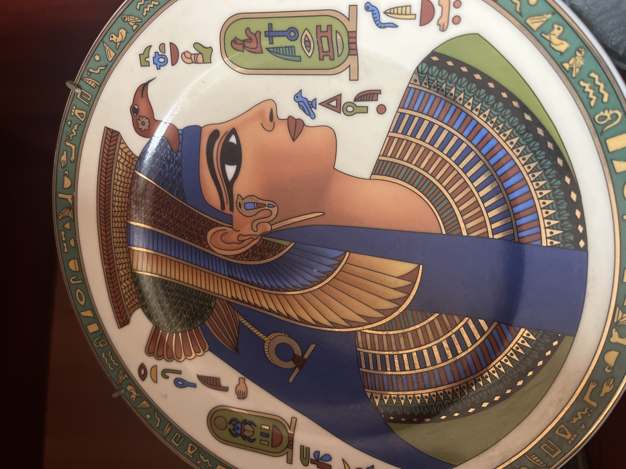 An ornate decorative plate, showcasing an Egyptian-themed design, is depicted. The central and dominant feature is a profile illustration of an ancient Egyptian royal figure, likely a pharaoh or queen, with distinct facial features, dark eye makeup, and a serene expression. This figure wears an elaborate headdress, possibly a nemes or royal wig, rendered in rich blues, golds, greens, and browns, with intricate striped patterns.
The plate itself is cream-colored, encircled by a wide decorative border in green, outlined in gold, and filled with various stylized ancient Egyptian hieroglyphs. Additional hieroglyphs are scattered around the figure's head, including a prominent cartouche above the forehead containing several symbols such as an ankh and a scarab beetle. Other individual hieroglyphs, resembling birds, hands, water, and various objects, are visible within the border and adjacent to the figure.
A small metallic wire hook is visible near the top left edge of the plate, suggesting it is either hanging or designed for display. The background behind the plate is dark and out of focus, indicating an indoor setting. The overall aesthetic evokes ancient Egyptian art and mythology, presented in a decorative, colorful style.