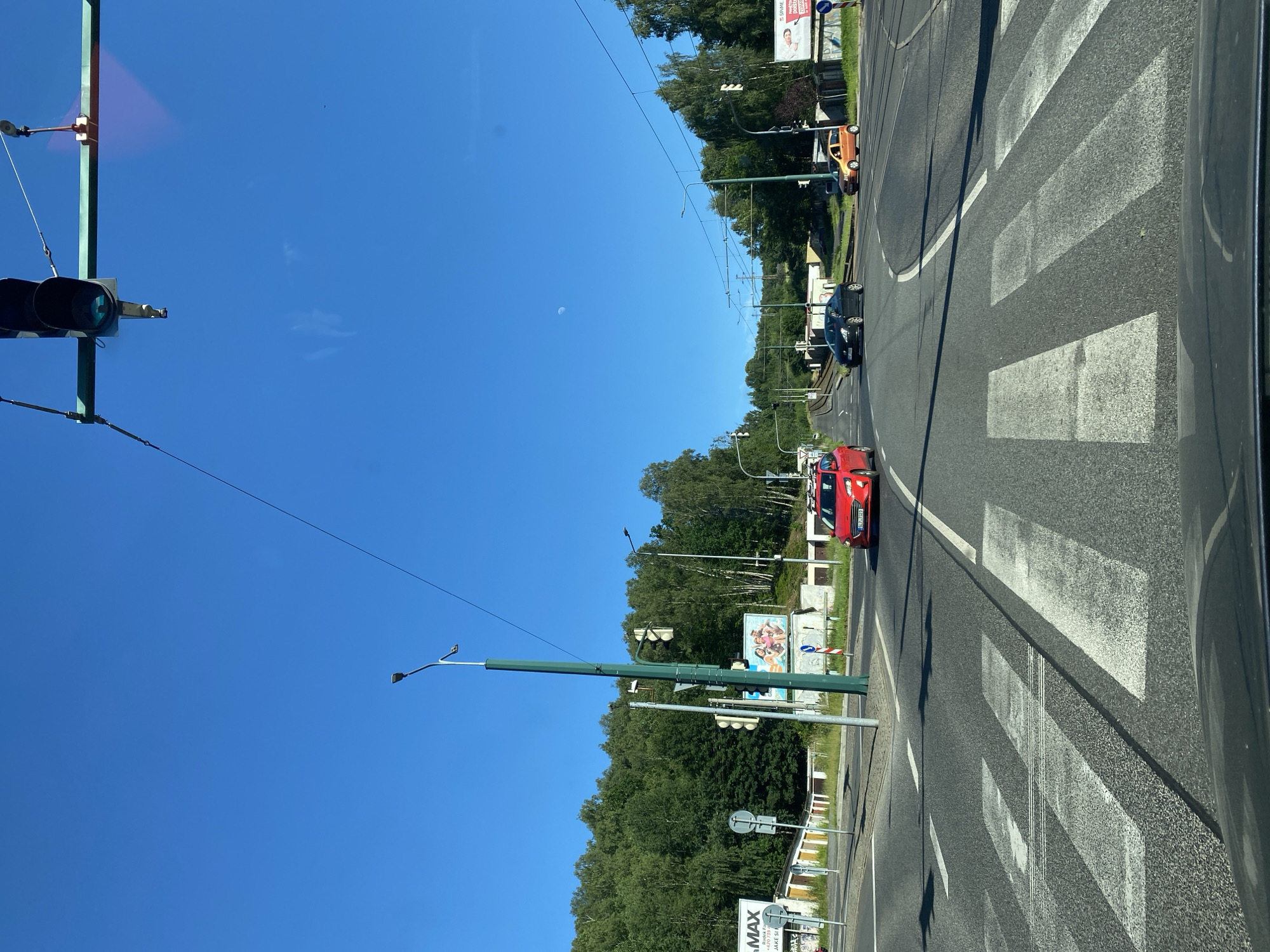 The image shows a street scene with a bright blue sky and lush green trees lining the road. A pedestrian crossing, marked by white stripes, is prominent in the foreground. Several cars are visible on the road, including a red car in the foreground, a black car in the middle distance, and an orange car further back. Traffic lights are present, suggesting an intersection or controlled crossing. The sun is shining, indicating it is daytime, and there are some light clouds in the sky, along with a visible sliver of the moon. Overhead wires, likely for an electric tram or trolleybus, are strung across the scene. There are also streetlights and billboards visible along the side of the road. One billboard appears to have people on it, possibly an advertisement. The scene gives the impression of a suburban or urban environment on a clear, sunny day.