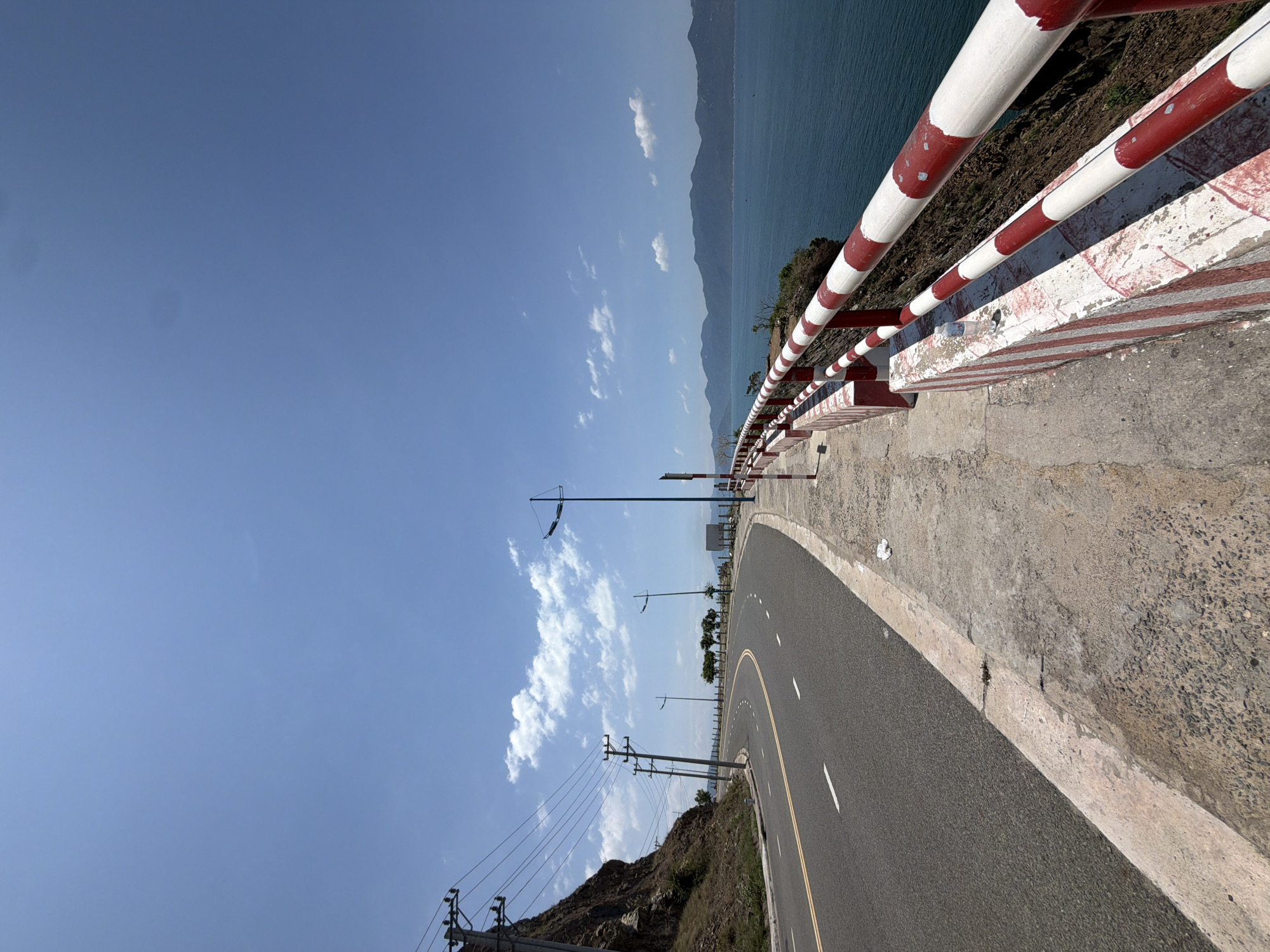 The image displays a curved coastal road situated in Bac Nha Trang Ward, Vietnam, under a clear blue sky with scattered white cumulus clouds. The asphalt road features double yellow center lines and dashed white lane markings, curving sharply to the left. A continuous white line delineates the right edge of the road, alongside a concrete sidewalk where a crumpled white wrapper is visible. Immediately adjacent to the sidewalk is a red and white striped concrete barrier, topped with matching red and white metal railings, overlooking the sea. A clear plastic water bottle rests on this barrier towards the right of the frame. The calm, deep blue sea stretches to the horizon, where distant mountain ranges are visible. Several small boats are present on the water in the distance. To the left of the road, a steep, rocky hillside rises, sparsely covered with vegetation. Multiple wooden utility poles with overhead wires run along the left side of the road, following the contour of the hillside. Several green-painted streetlights are positioned along the road's curvature. The scene is brightly illuminated by natural sunlight. No individuals or vehicles are present within the captured area.