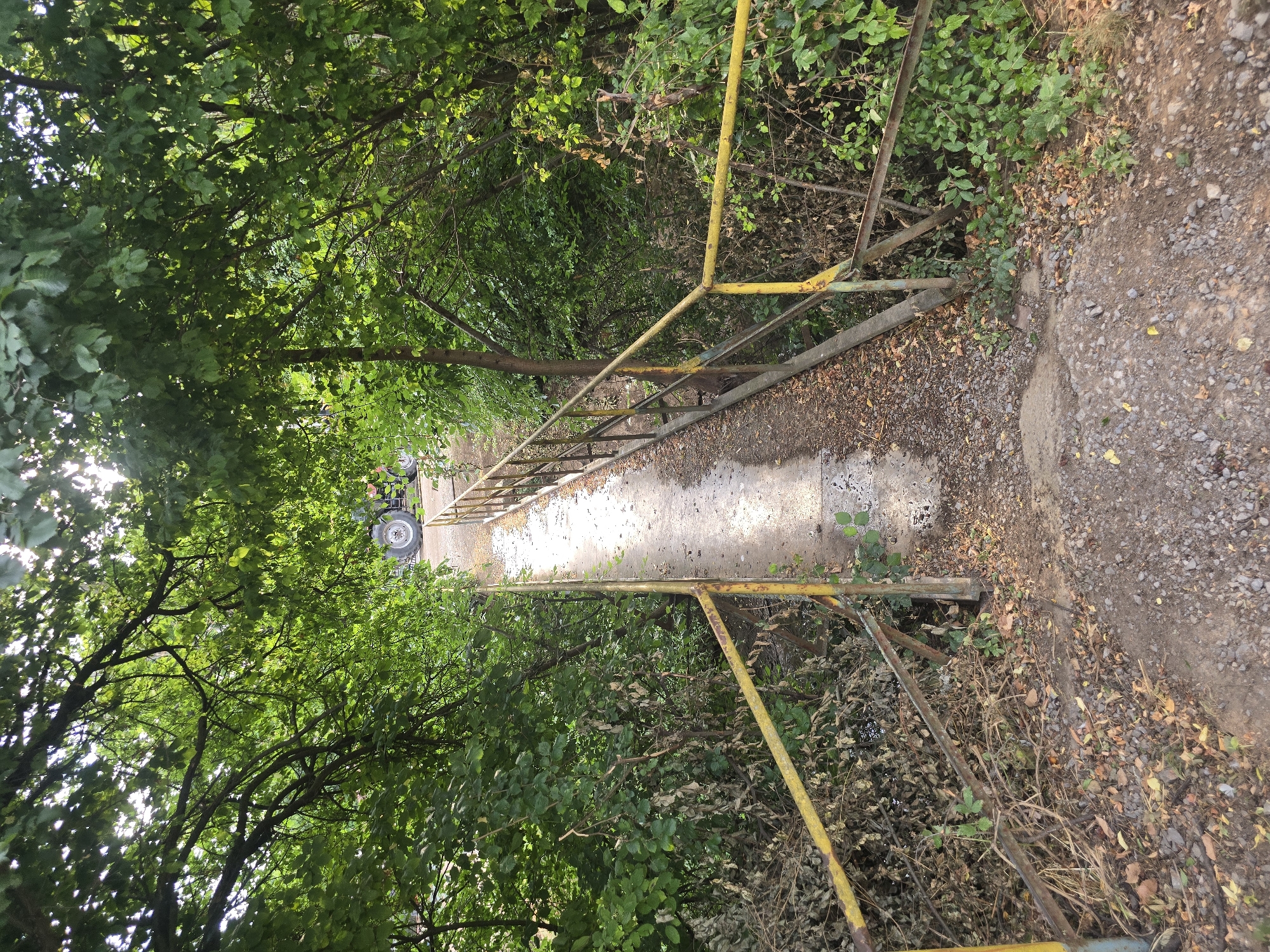 An elevated perspective looks down a narrow, concrete bridge overhung with lush green trees. The bridge has a yellow metal railing on both sides, which appears aged and somewhat rusty in places. The bridge surface is worn and uneven, with scattered gravel and fallen leaves. Beyond the bridge, a large tractor with prominent tires is partially visible, suggesting an agricultural or industrial setting. The surrounding vegetation is dense, with a variety of trees and bushes creating a somewhat secluded atmosphere. The lighting suggests it is daytime, with sunlight filtering through the leaves.