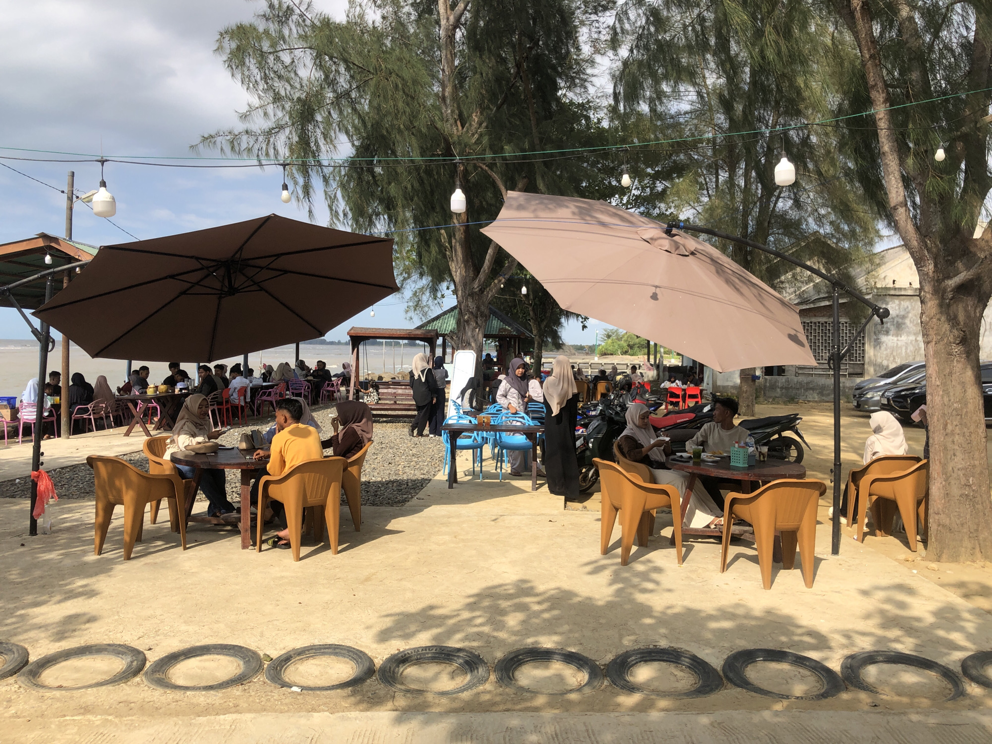 The image captures a vibrant outdoor dining scene in Lancok, Indonesia. The setting is a sandy area dotted with tables and chairs, partially shaded by large brown umbrellas and the foliage of mature trees. People are seated at the tables, engaged in conversation and dining. Some wear traditional Muslim head coverings. Parked vehicles are visible in the background, suggesting a location near a road. The ground has a decorative border made of tires. Overhead, string lights hang from the trees. A beach and water feature are visible in the background.