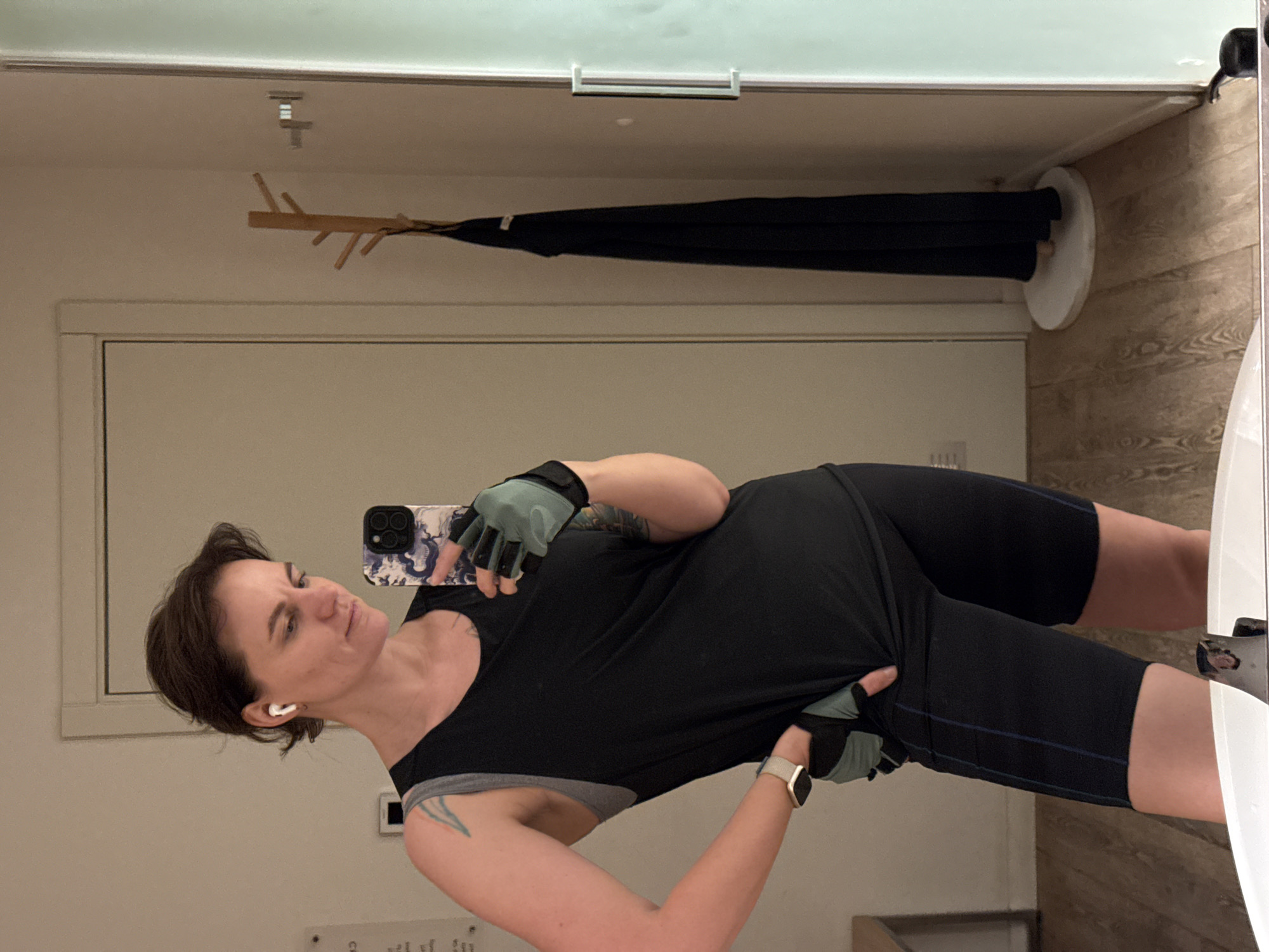 A woman with short brown hair is taking a mirror selfie in a gym or workout area. She is wearing a black tank top over a gray sports bra, black athletic shorts with a blue stripe, and workout gloves. An Apple Watch is visible on her left wrist. She is holding a smartphone with a patterned case. Behind her is a white door, a wooden coat rack with a black garment hanging on it, and a wall with light beige vertical paneling. The location is assumed to be a gym or workout area based on the woman's attire and the setting. The city of Boreti, Montenegro is not mentioned in the image.