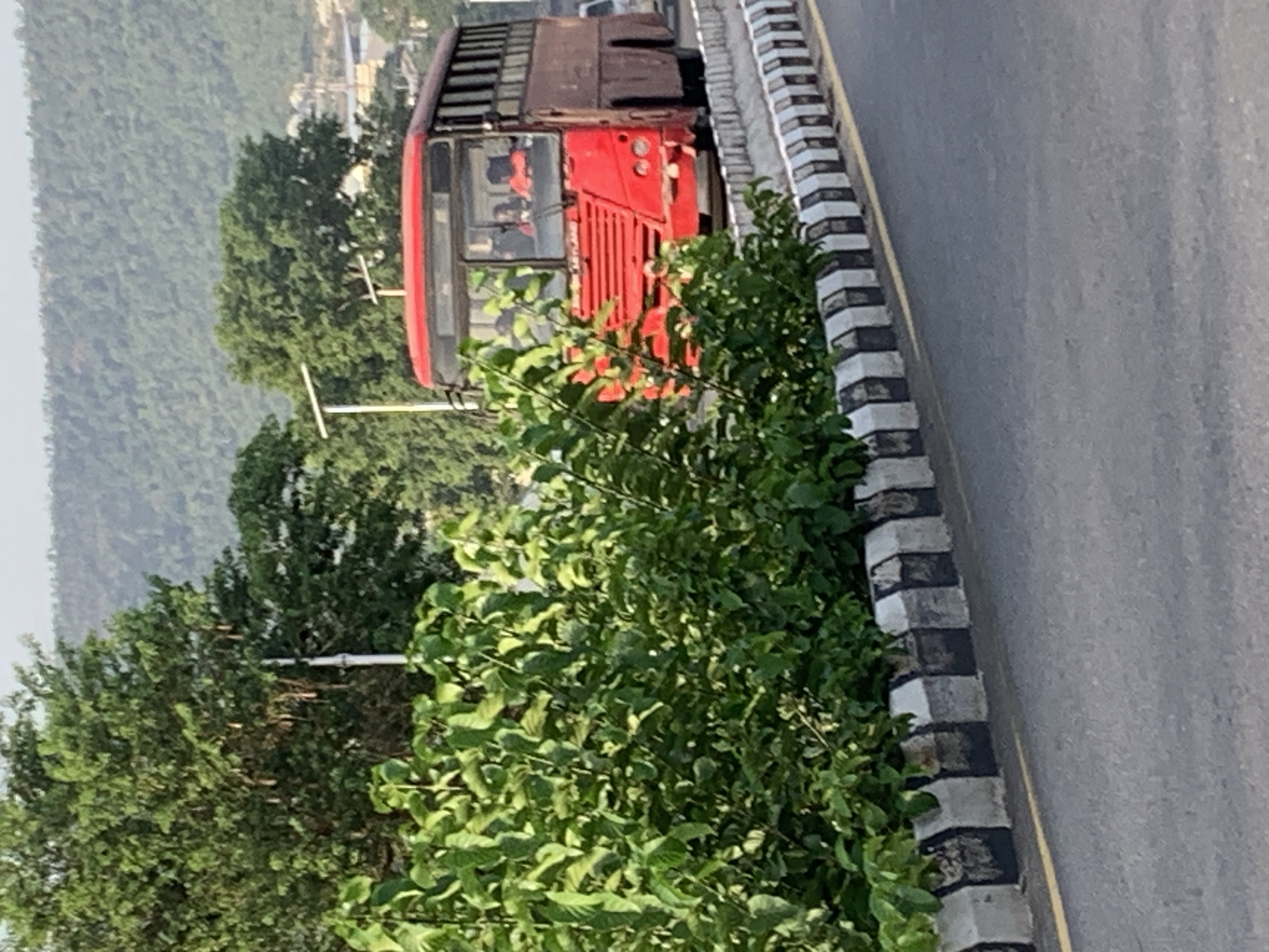 A medium shot of a red bus with a brown luggage rack on top driving along a road in Zango-ndaji, Nigeria. The bus is partially obscured by leafy green vegetation in the foreground. The road has a black and white curb along its edge. In the background are green trees and a hazy hillside.