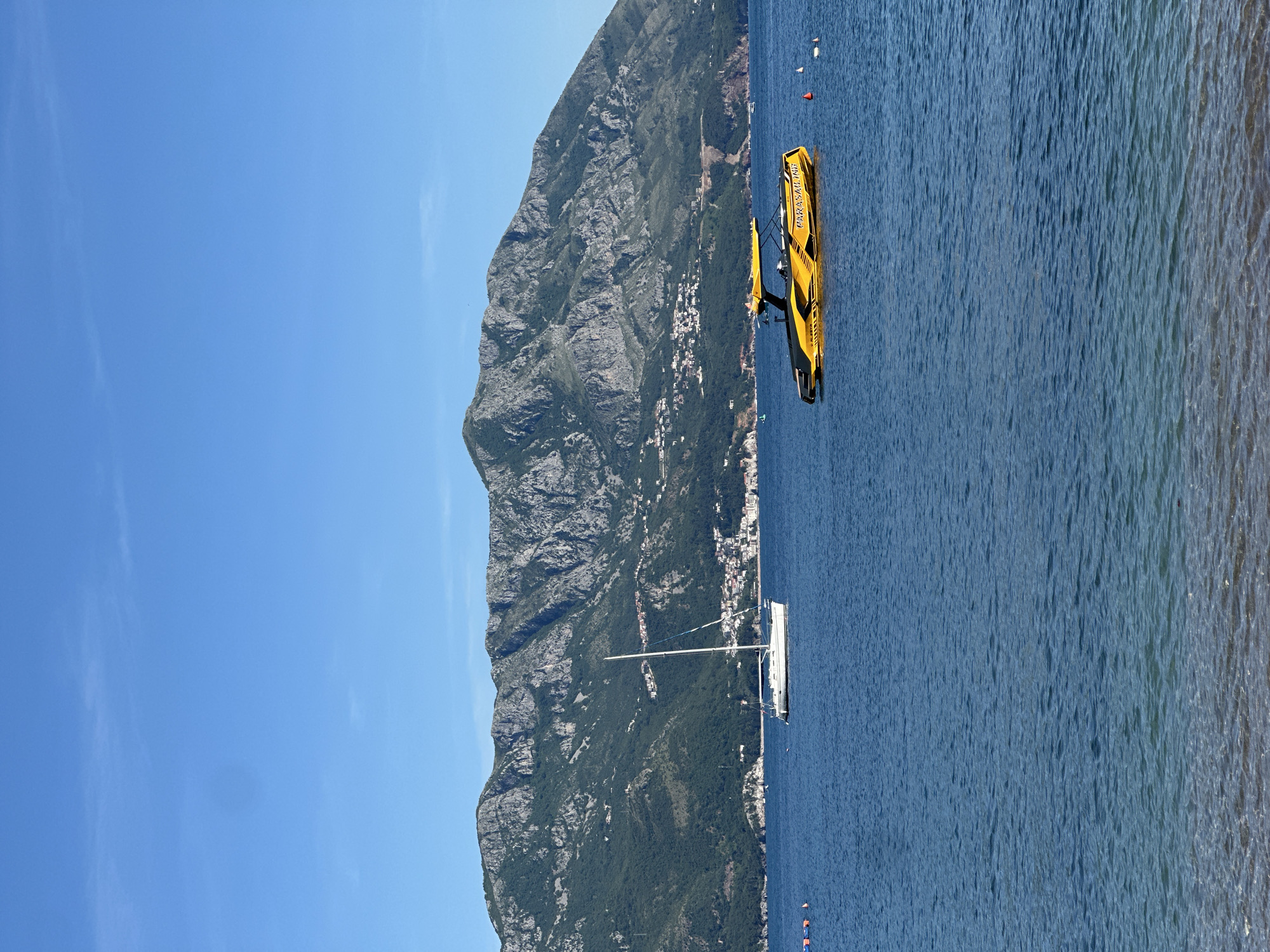Captured in Budva, Montenegro, the image presents a scene with diverse elements. Dominating the left of the frame is a craggy mountain range, blanketed in greenery. Adjacent to the mountains, a scattering of buildings hints at a coastal settlement.
On the right, the expanse of the sea is dotted with floating objects that appear to be navigational markers. A yellow parasailing boat is visible mid-frame, and a white sailing boat stands further in the distance. The rightmost edge of the image shows the coastline with its sandy beach. The sky above is cloudless, a clear azure blue. The location is characterized by coastal mountains, the sea, and boats, suggesting a maritime recreation area.