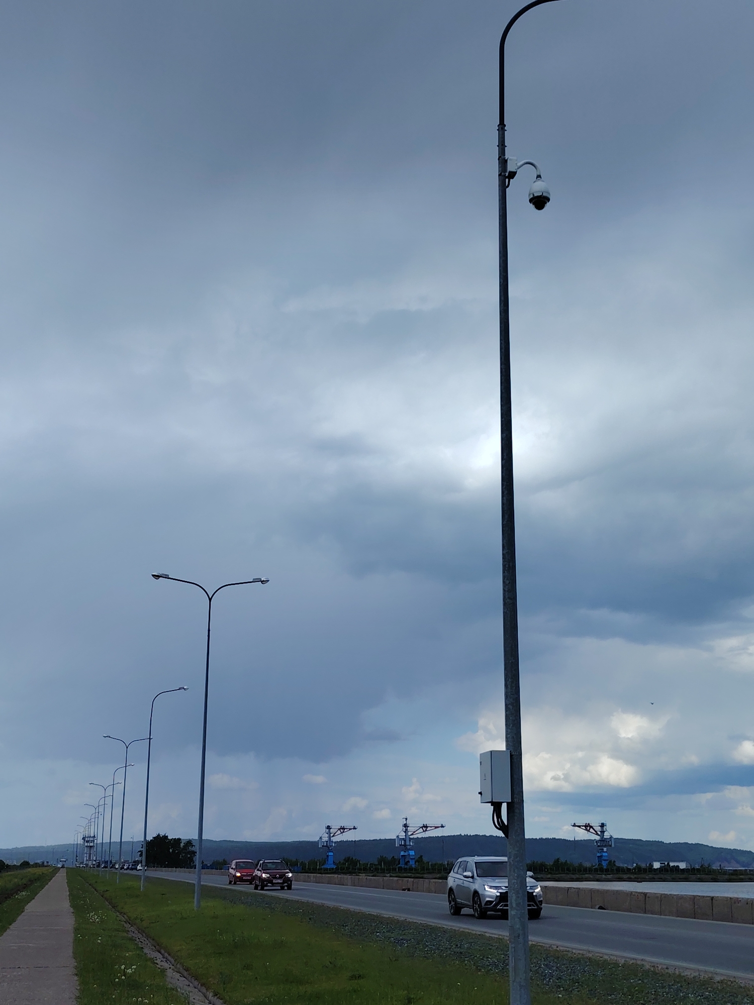 The image depicts a road leading through the city of Chaikovsky, Russia. Along the roadside, there are several evenly spaced lampposts. One lamppost closer to the foreground also has a surveillance camera. There are vehicles visible on the road: two cars passing in opposite directions. In the background, cranes can be seen near the river. The sky is cloudy, suggesting an overcast or potentially stormy weather condition. A sidewalk runs along one side of the road.
